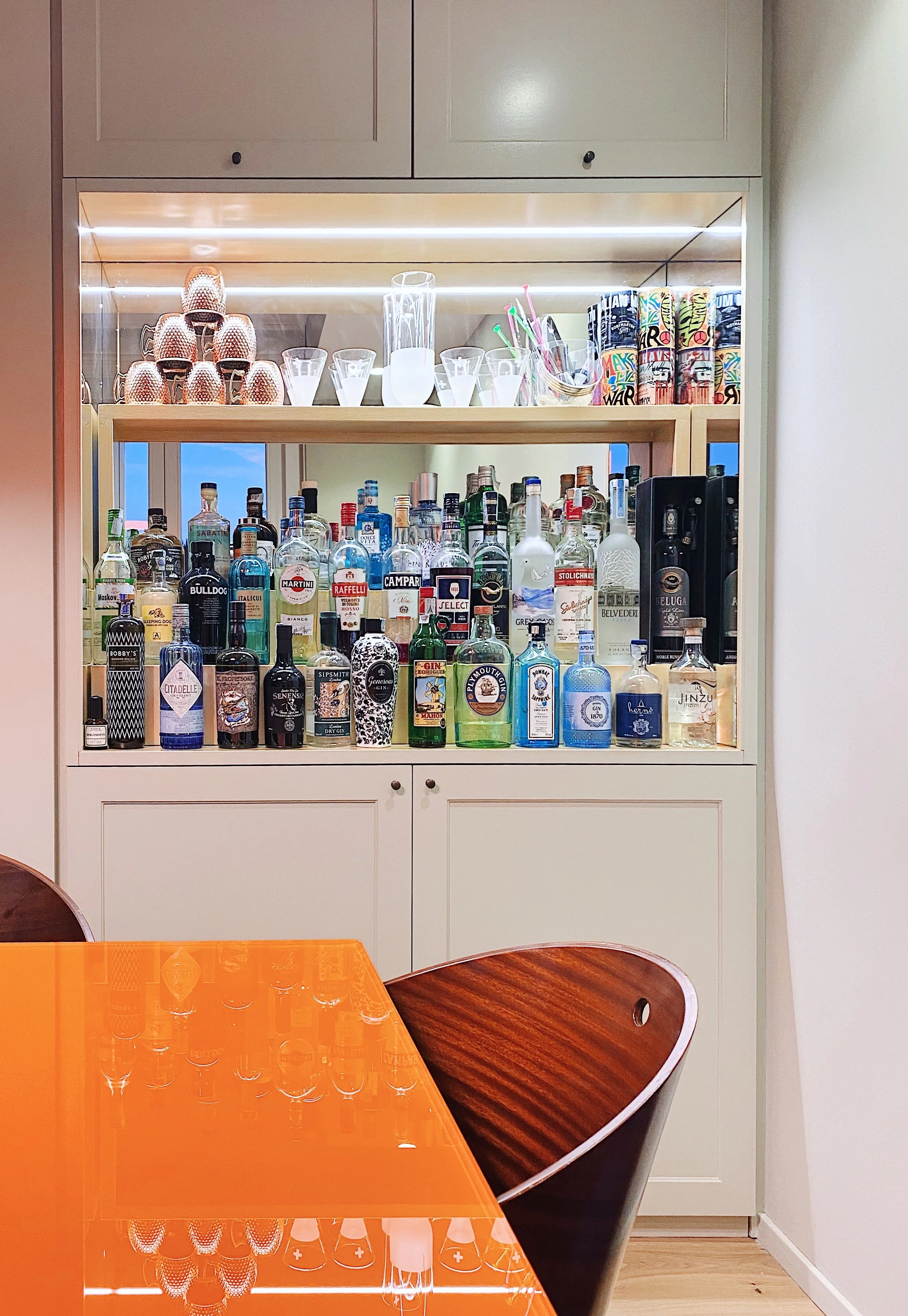Stanza con bar con bottiglie di alcolici, bicchieri, e oggetti decorativi, vicino a tavolo con superficie riflettente arancione.