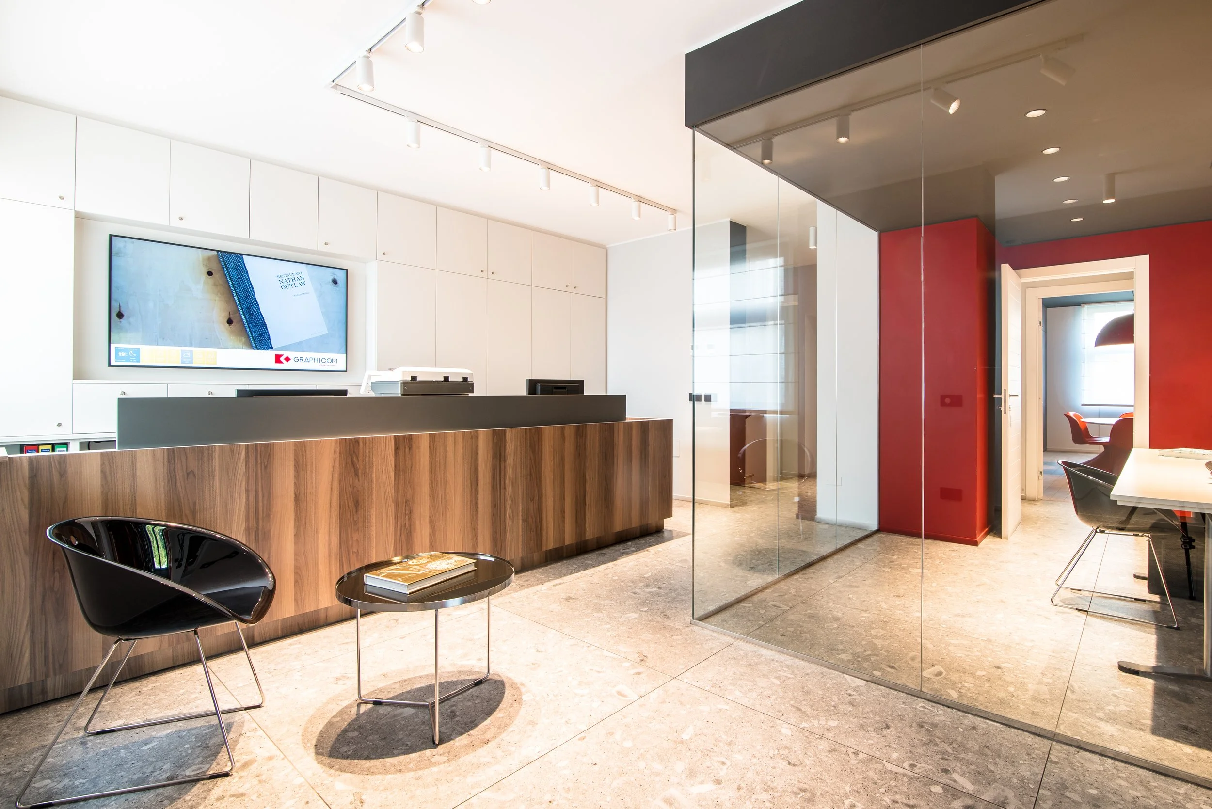 Reception room with a wooden front desk, a black chair and a small round table with a magazine, white walls, a large monitor on the wall, glass partitions, red accent wall, and a room with dining table and chairs visible through an open door.