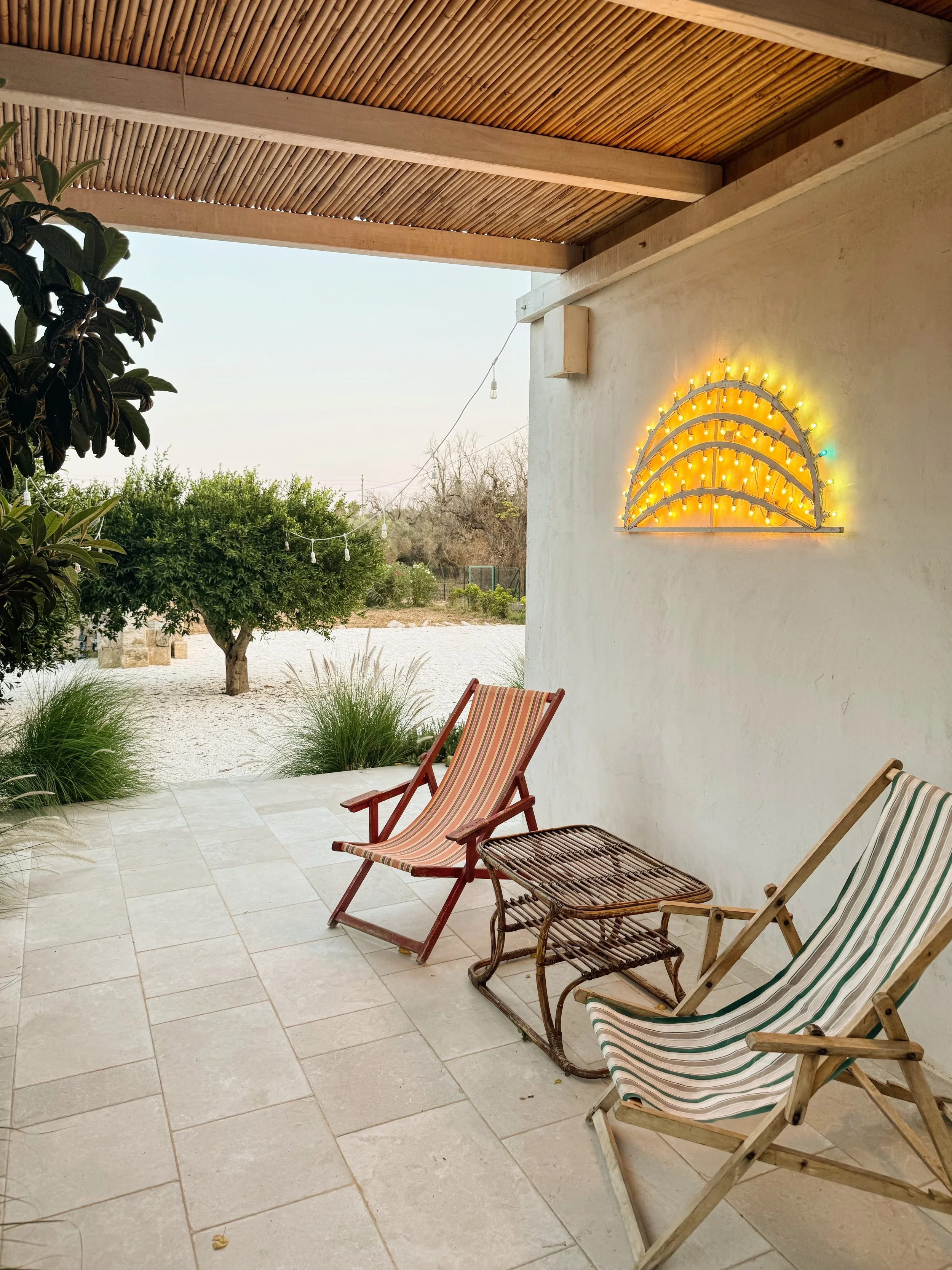 Veranda con due sedie a sdraio con tessuto a righe, tavolino in vimini, piante e alberi sullo sfondo, decorazione luminosa sulla parete.