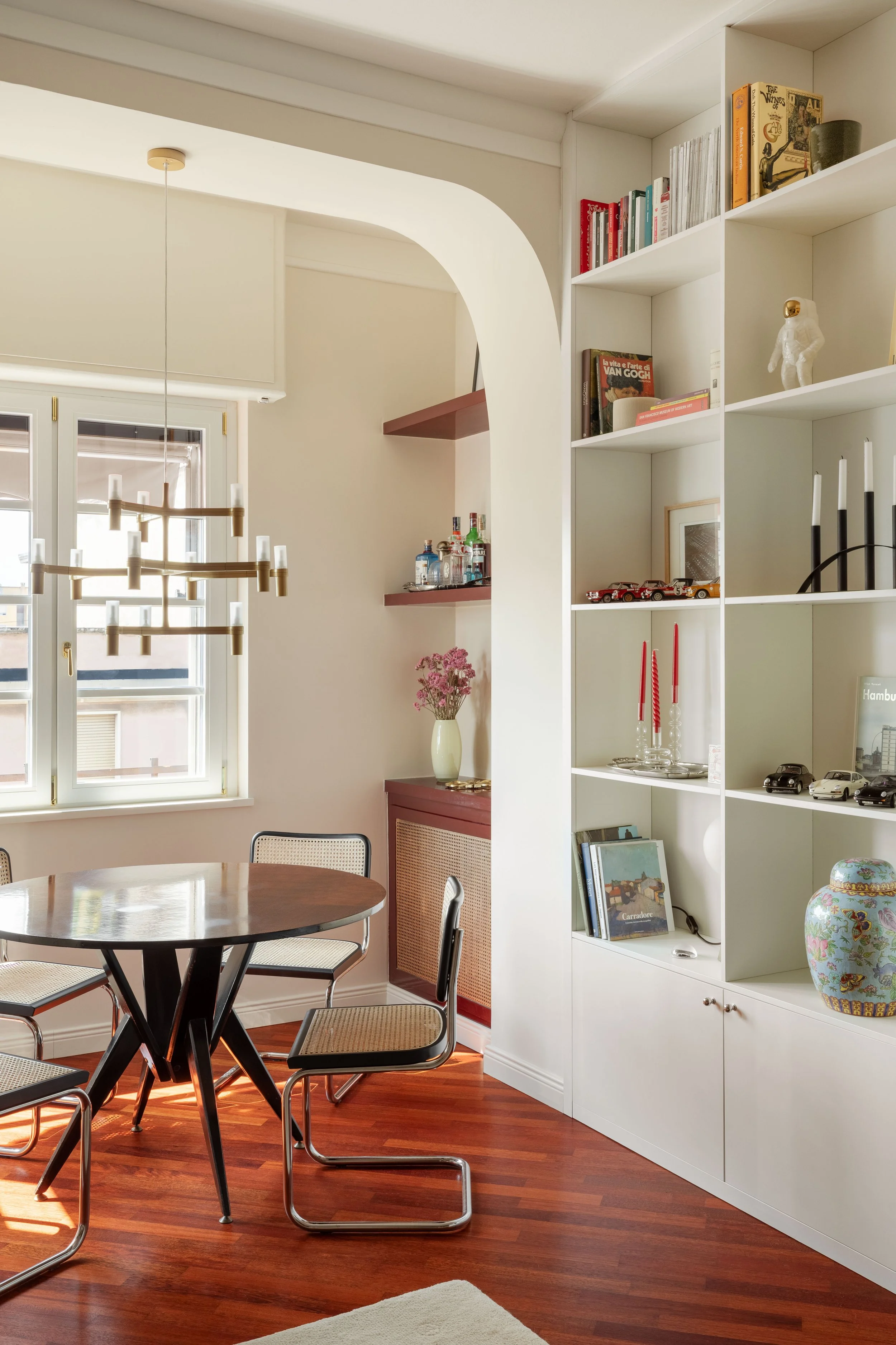 Interior di un soggiorno con pareti chiare, grande finestra con tende bianche, lampadario moderno, tavolo rotondo con sedie, scaffale con libri, decorazioni e modellini di automobili, pianta con fiori rosa, pavimento di legno.