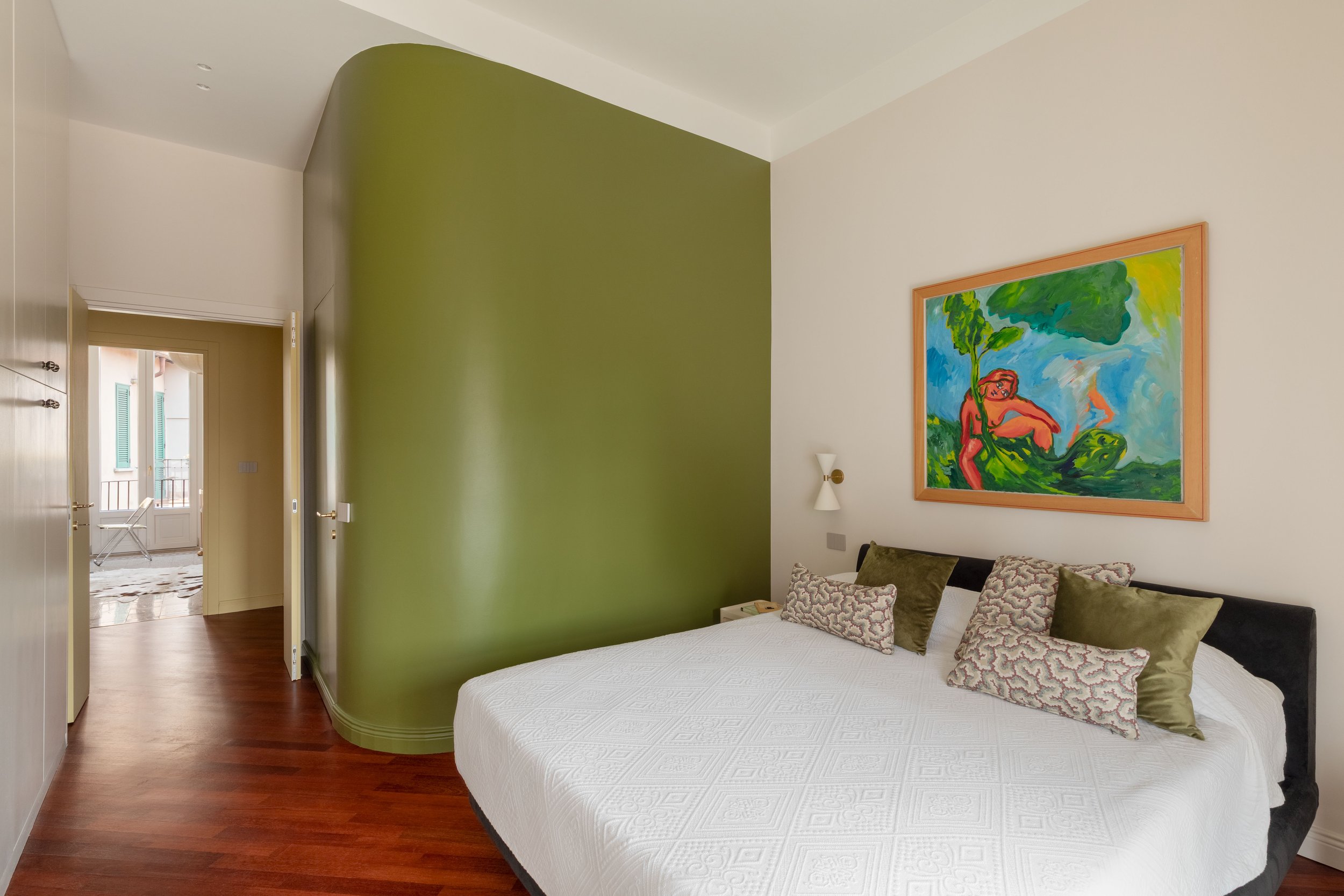 Interno di una stanza con pareti color crema e verde, decorata con un quadro colorato e cuscini su un letto. Una porta aperta conduce a un balcone con sedie.