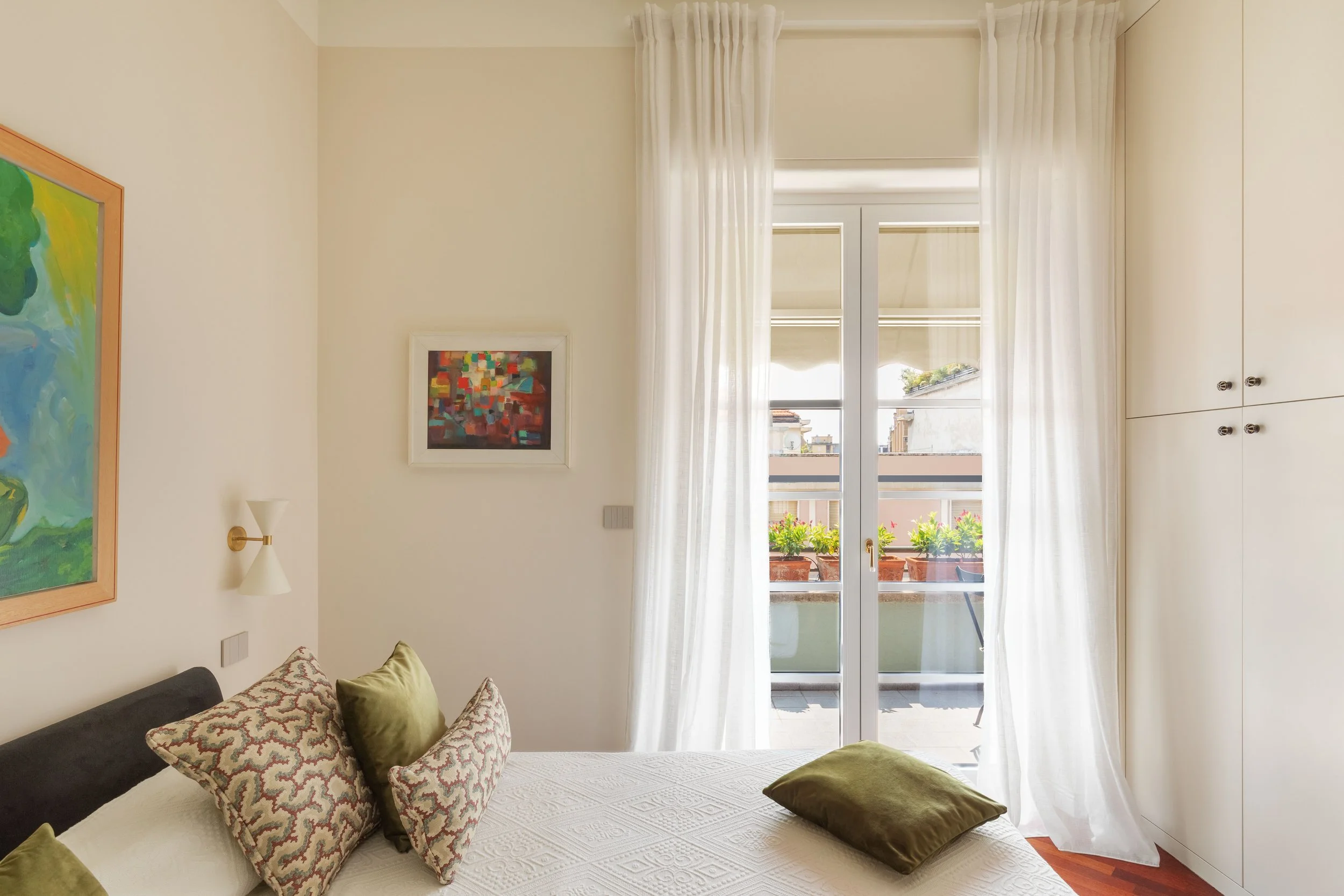 Camera da letto con pareti chiare, presenta due quadri, tende bianche trasparenti su una finestra grande che si affaccia su un balcone con piante. Sul letto ci sono due cuscini verdi e due con motivi rossi e beige.