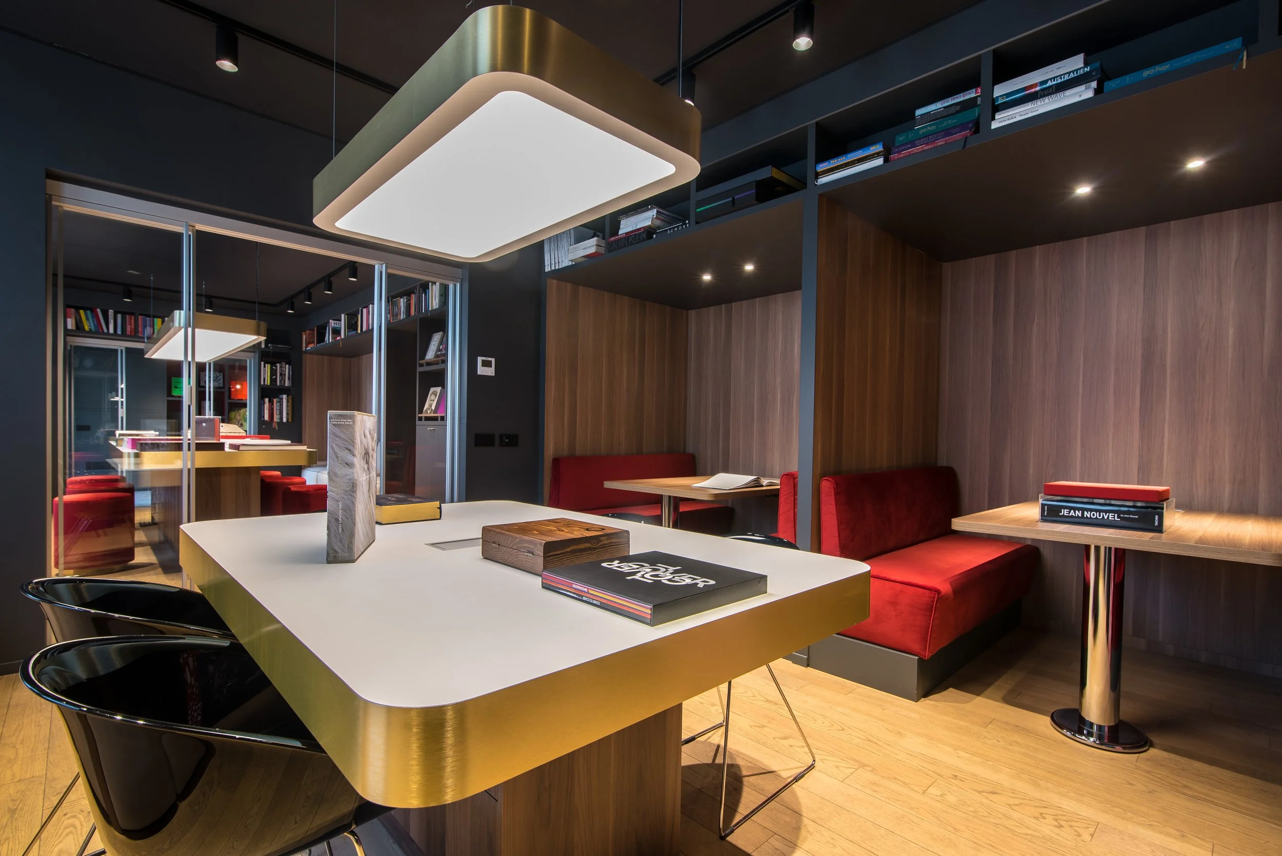 Interno di una sala moderna con tavoli, sedie nere, divanetti rossi, librerie con libri e illuminazione a soffitto.
