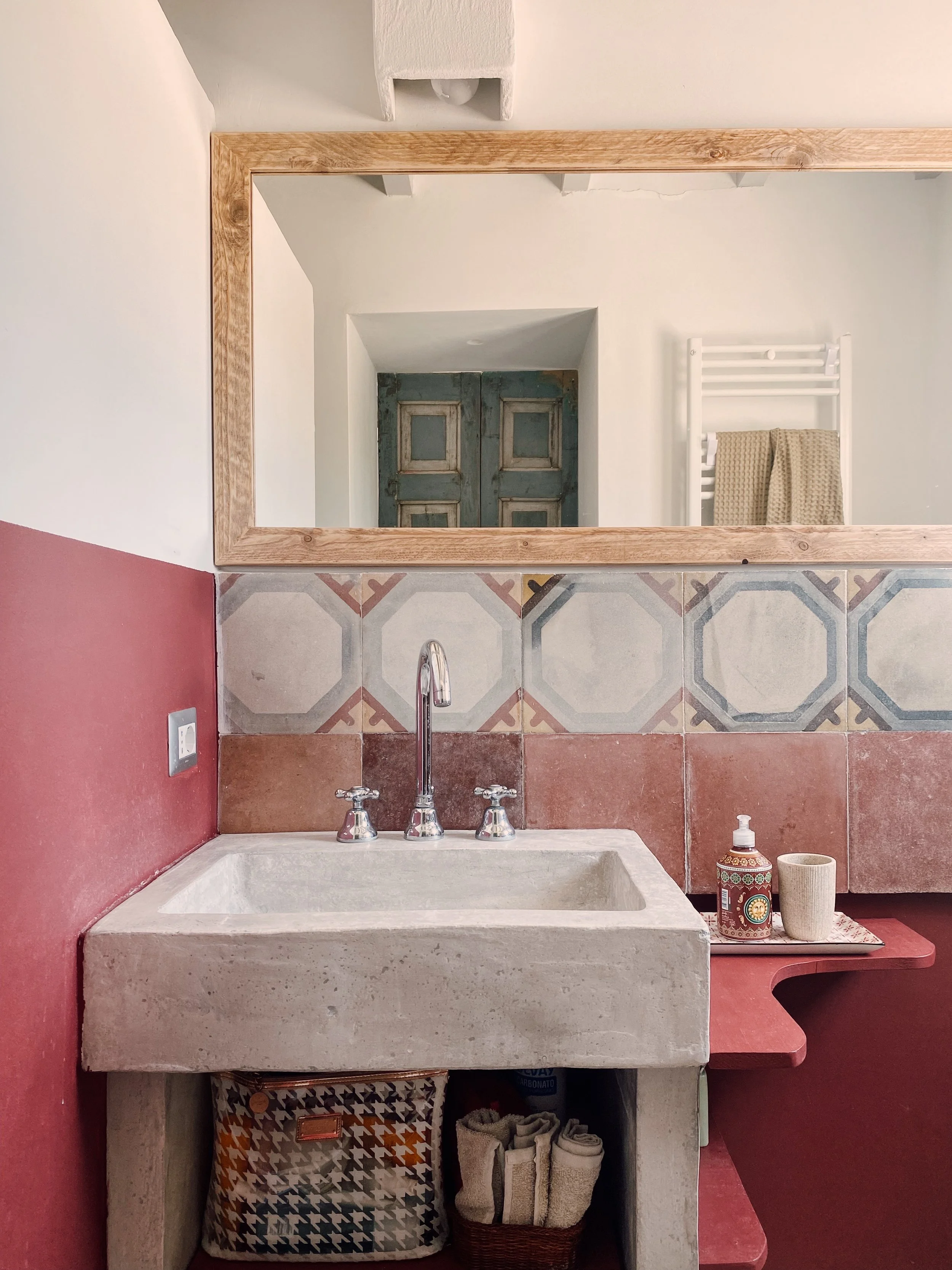 Lavandino in pietra con rubinetto a due manopole, specchio grande con cornice di legno riflesso in una parete bianca, pareti dipinte di rosso, accessori di bagno come sapone e bicchieri, scaffale sotto il lavabo con ceste di asciugamani.