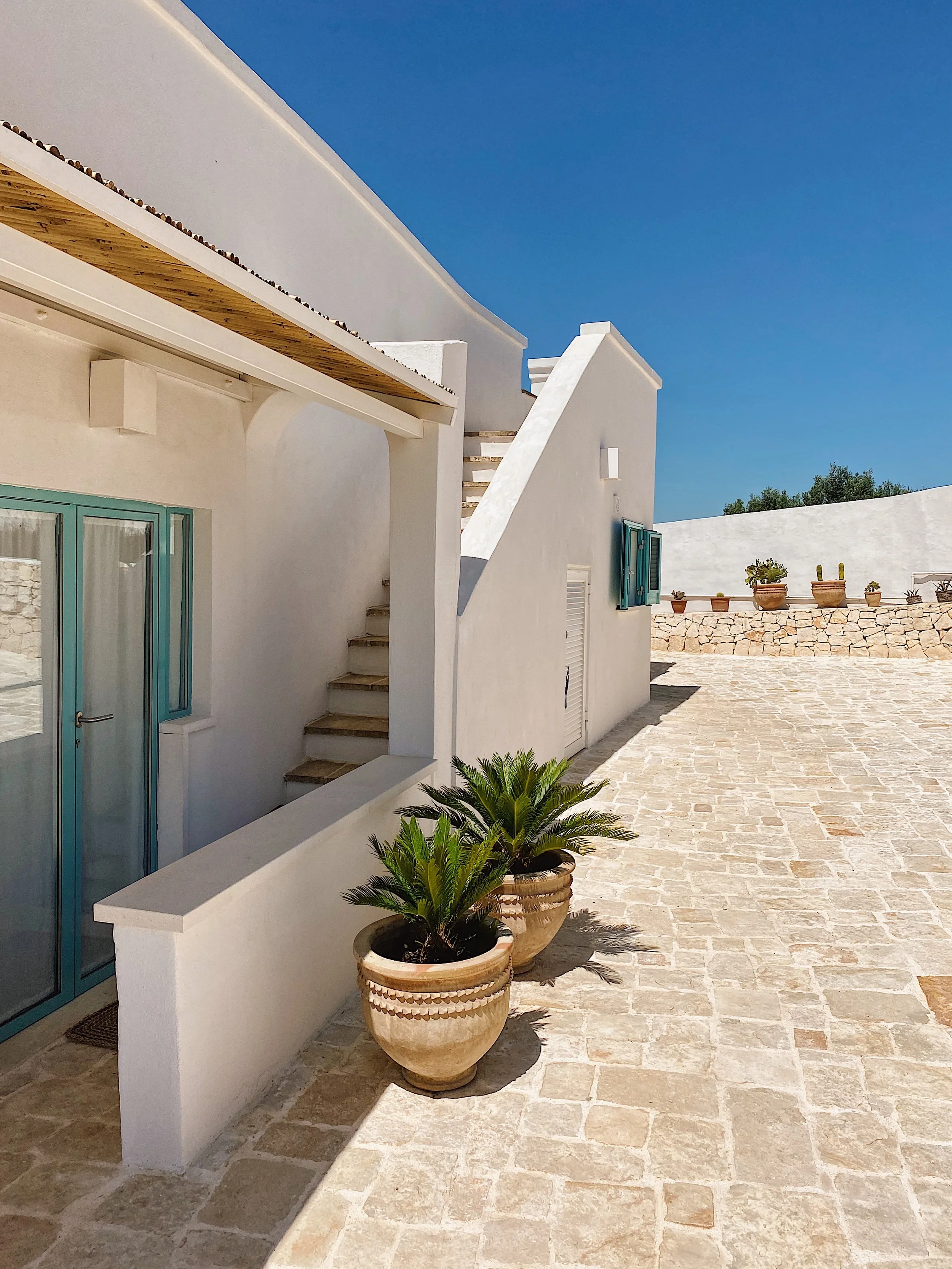 Cortile con pavimento in pietra e piante in grandi vasi di terracotta, case bianche con finestre e scale.