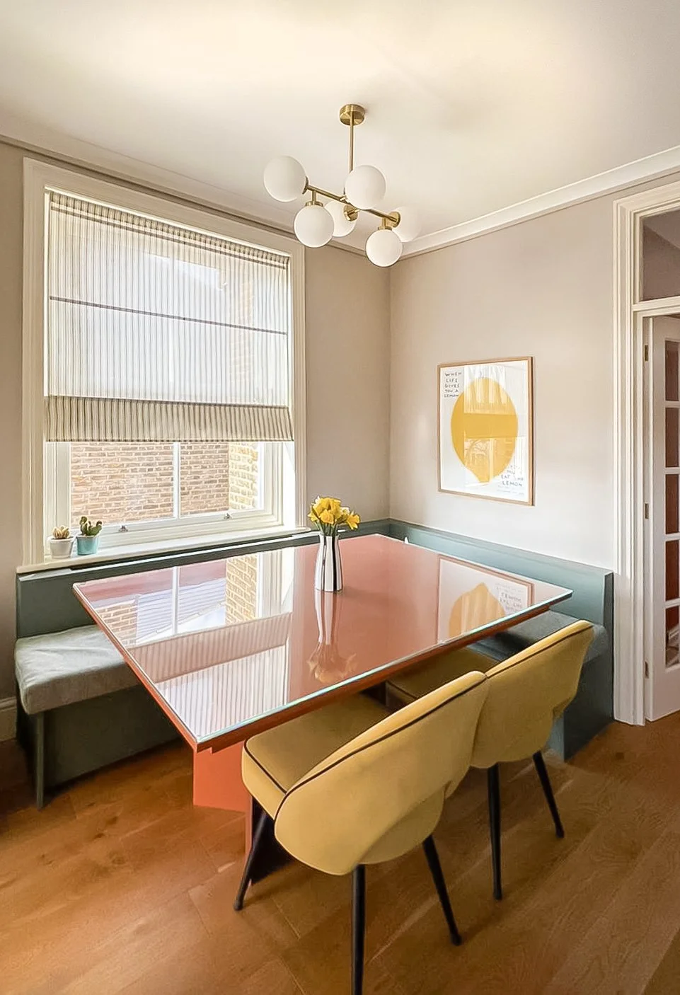 Una stanza da pranzo con finestra, tavolo rosa con tovagliolo, sedie gialle e decorazioni floreali, quadro astratto giallo e bianco, lampadario moderno con sfera. 