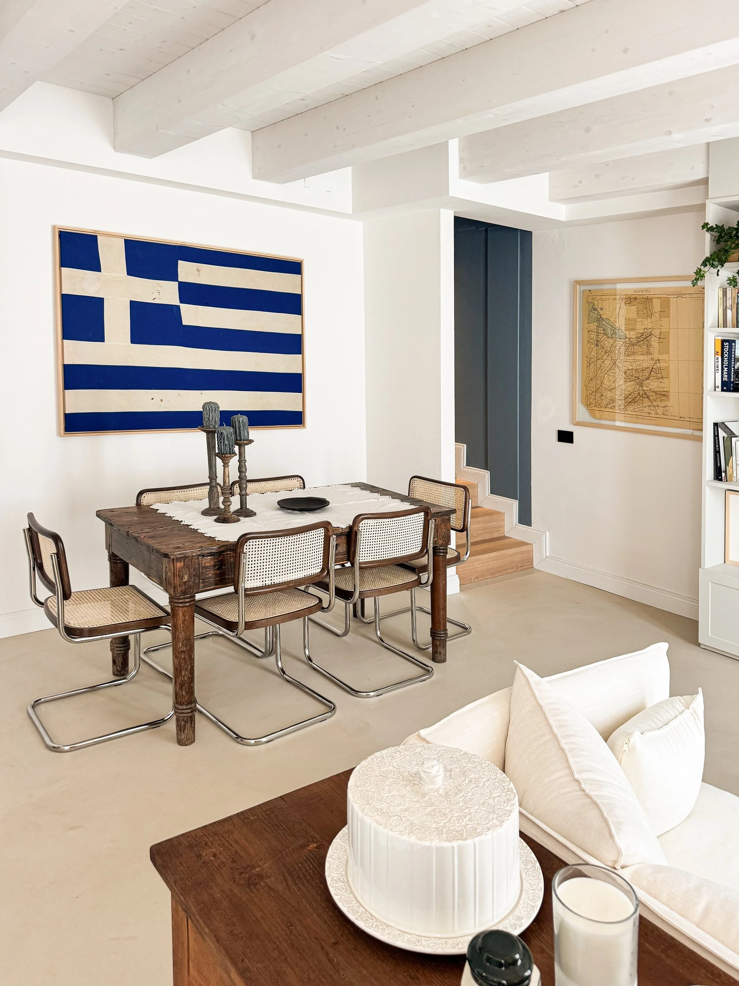 Interno di una casa con pareti bianche e soffitto con travi di legno chiaro. Sul muro di sinistra, un quadro con la bandiera della Grecia. Tavolo in legno con quattro sedie, al centro candelabro con tre candele. Sul tavolo, una torta bianca decorata 
