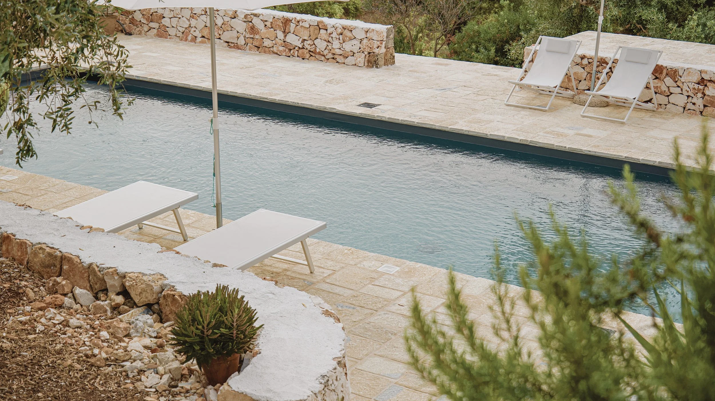Piscina all'aperto con sedie a sdraio bianche e ombrellone, circondata da pietre e piante.