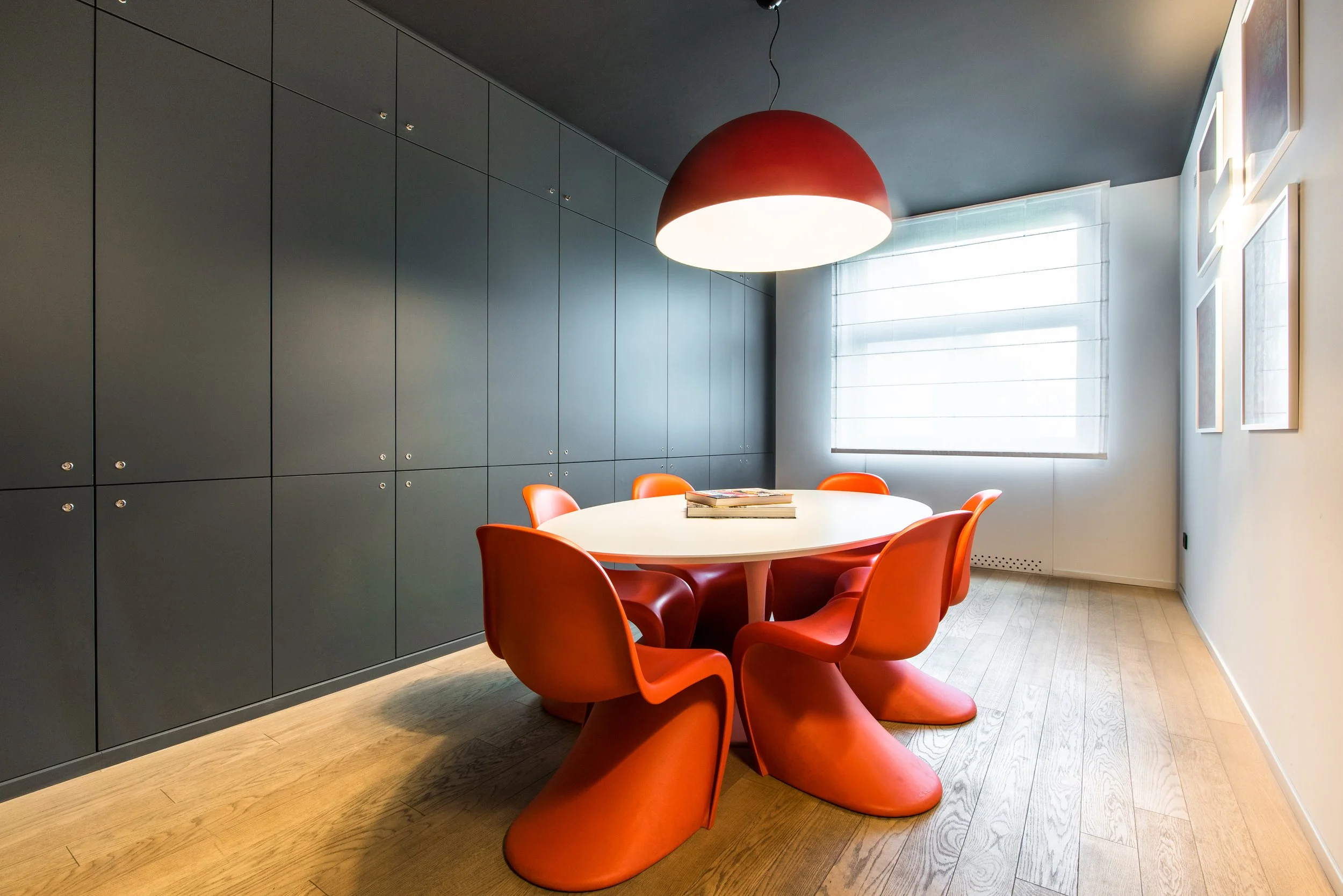 Sala riunioni con tavolo rotondo bianco, sedie arancioni futuristiche, grande lampada rossa e bianca, armadietti blu scuro, pareti chiare con quadri e finestra con tenda bianca.