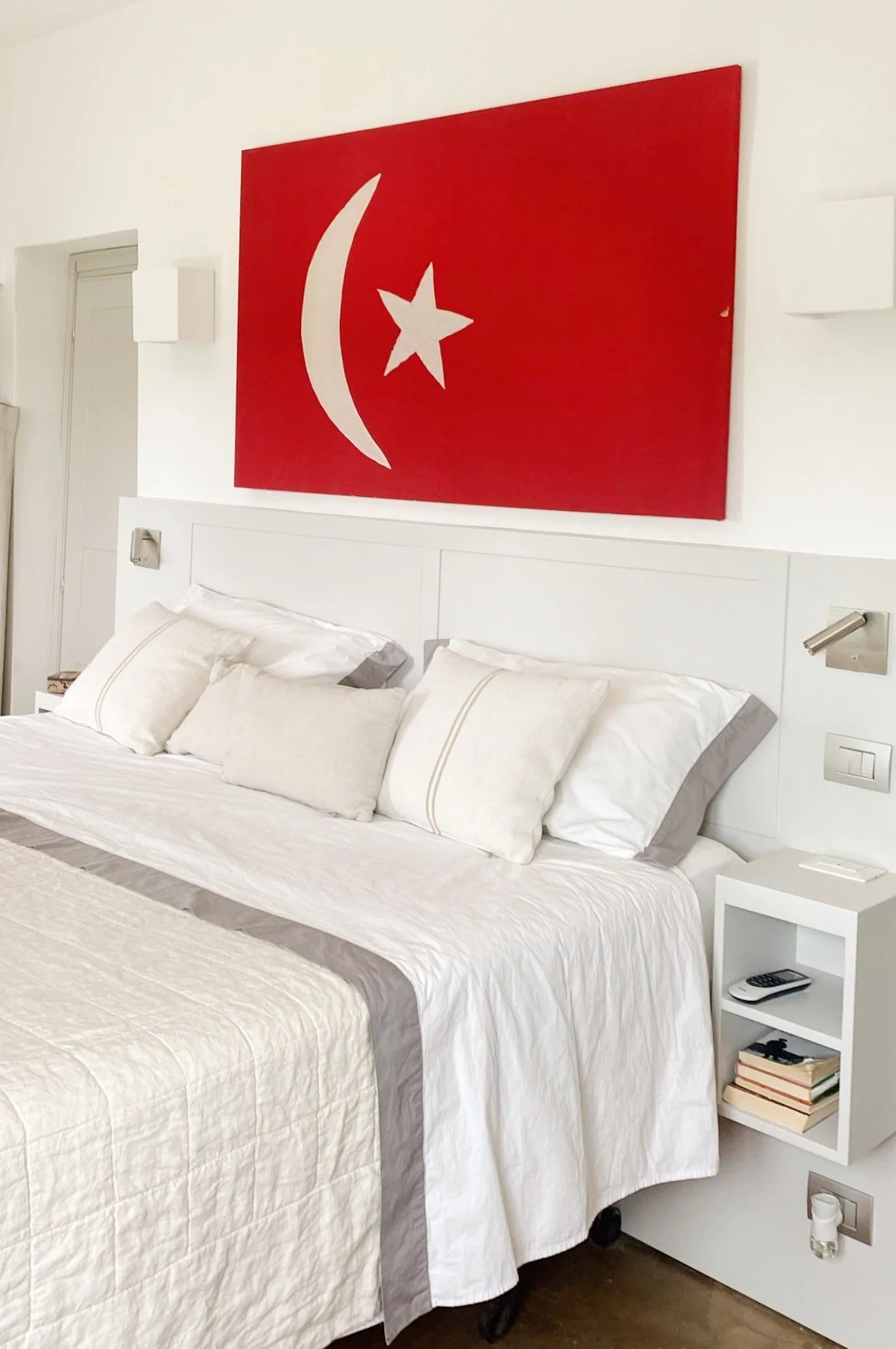 Letto con cuscini bianchi, sfondo di una parete bianca con un quadro rosso con simbolo della luna, stella e mezzaluna di colore bianco.