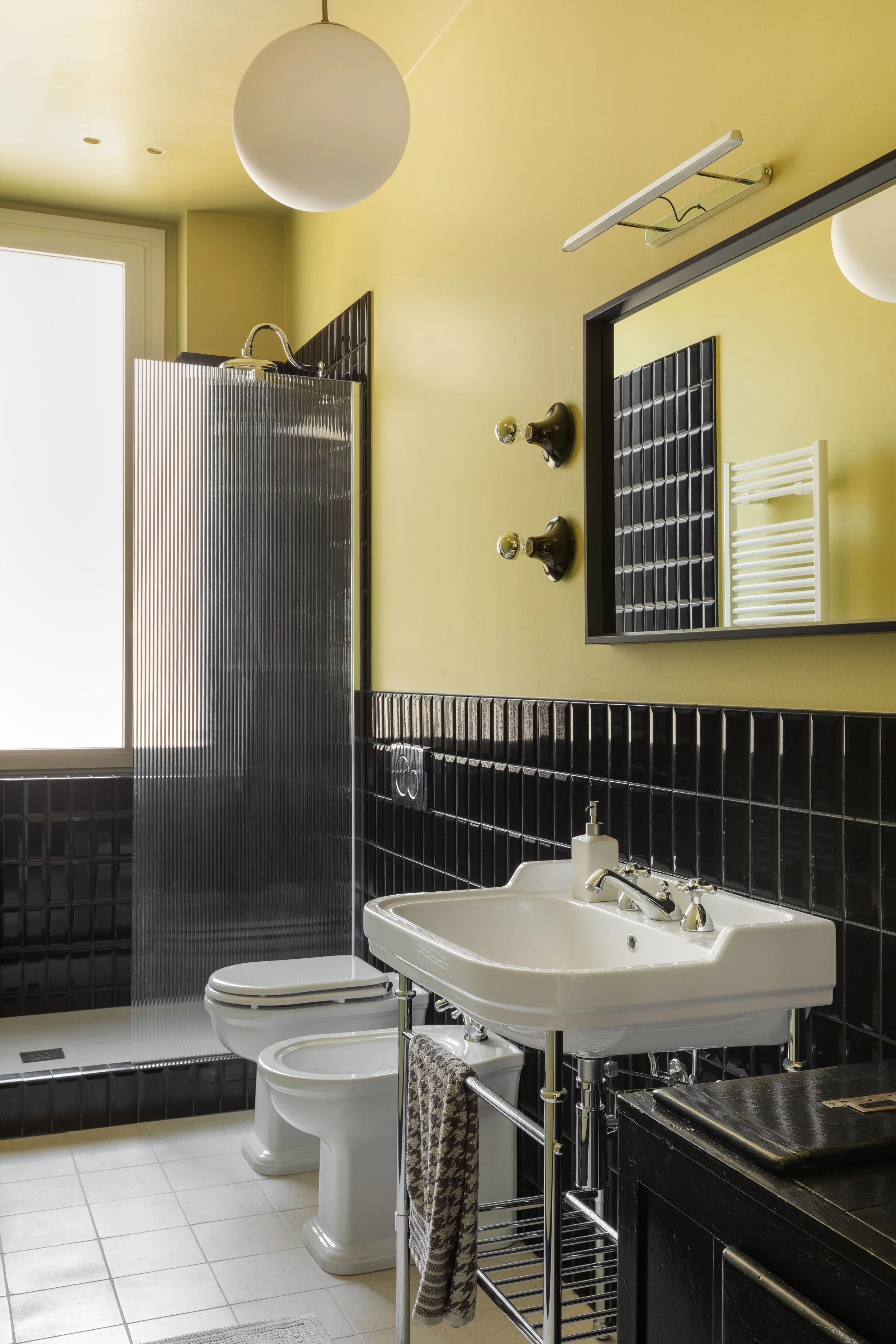 Bagno moderno con pareti gialle, rivestimenti in piastrelle nere, lavabo bianco con accessori in acciaio e specchio grande. L'illuminazione a soffitto e il termosifone bianco completano l'ambiente.
