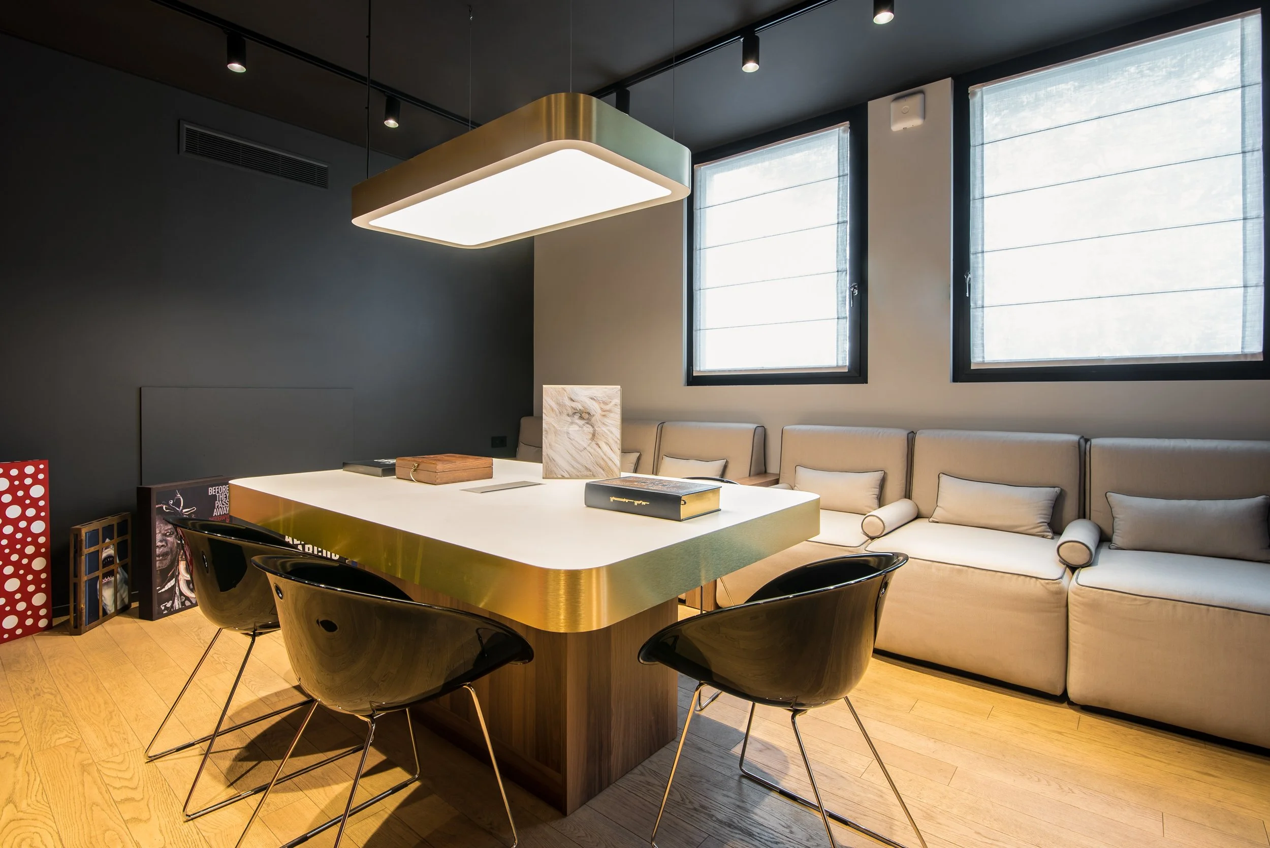 Sala riunioni moderna con grande tavolo bianco, sedie nere intorno, divano beige lungo il muro, due finestre con tende e lampada rettangolare sospesa sopra il tavolo.