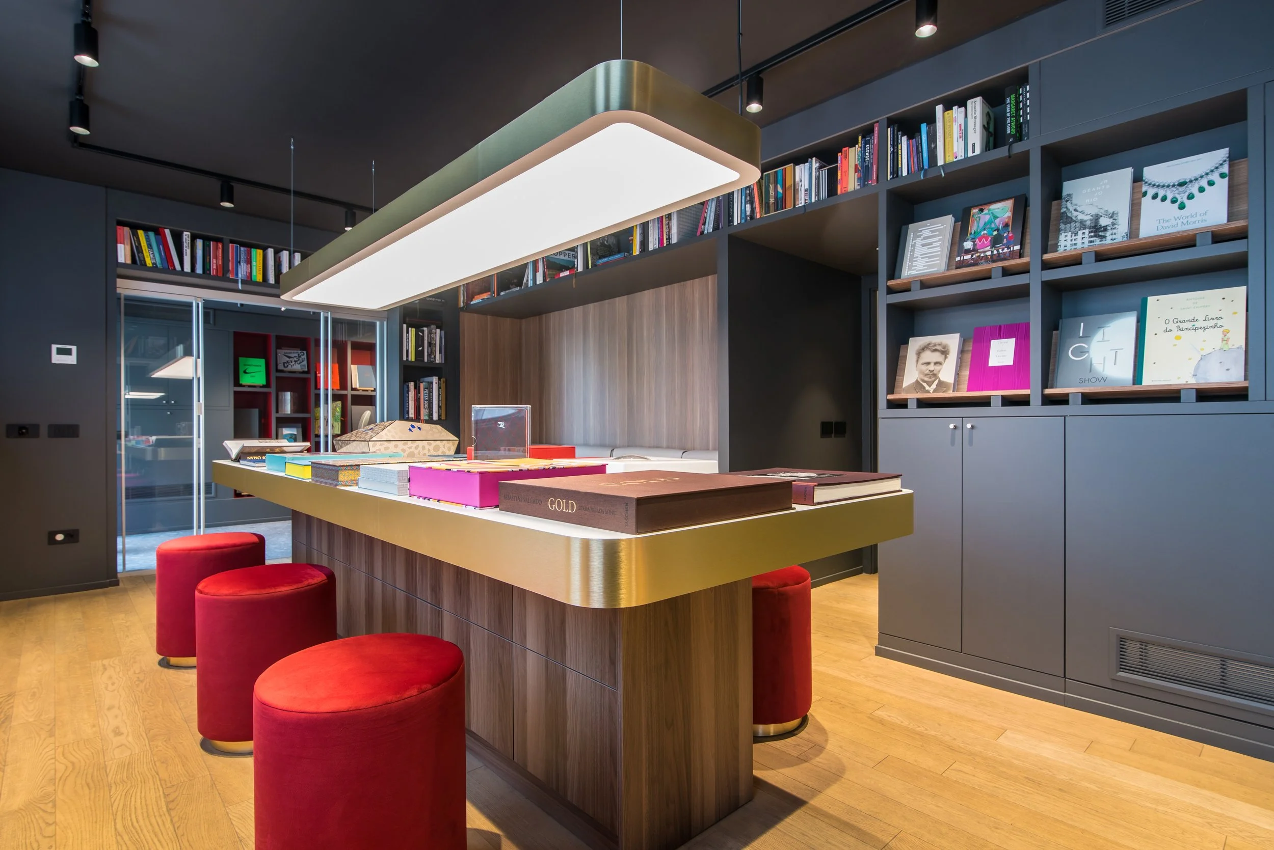 Interno di una libreria o biblioteca con scaffali di libri colorati e un tavolo centrale con libri e oggetti, illuminato da lampade a soffitto e sedie rosse in stile moderno.