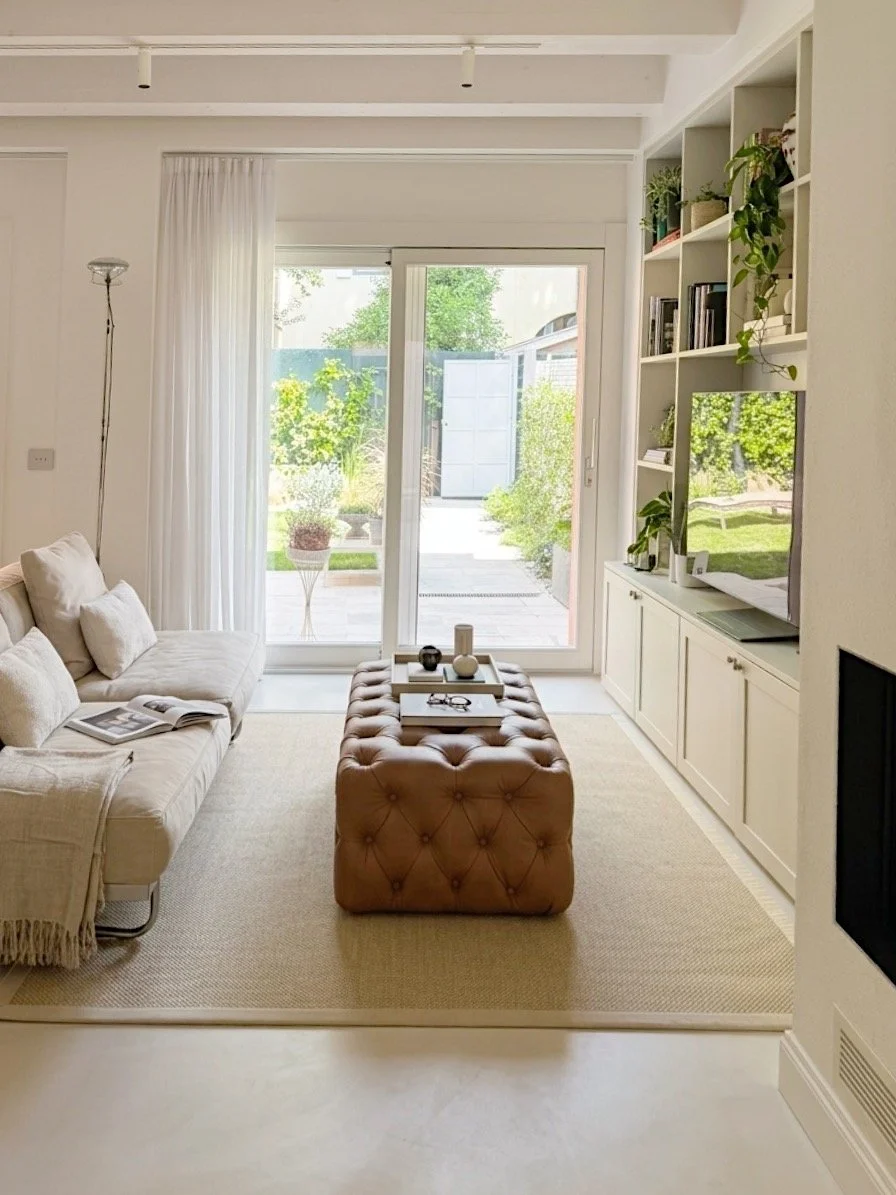 Soggiorno luminoso con divano bianco, pouf marrone, libreria con piante e televisore, grande porta finestra con tende che apre su un giardino verde.