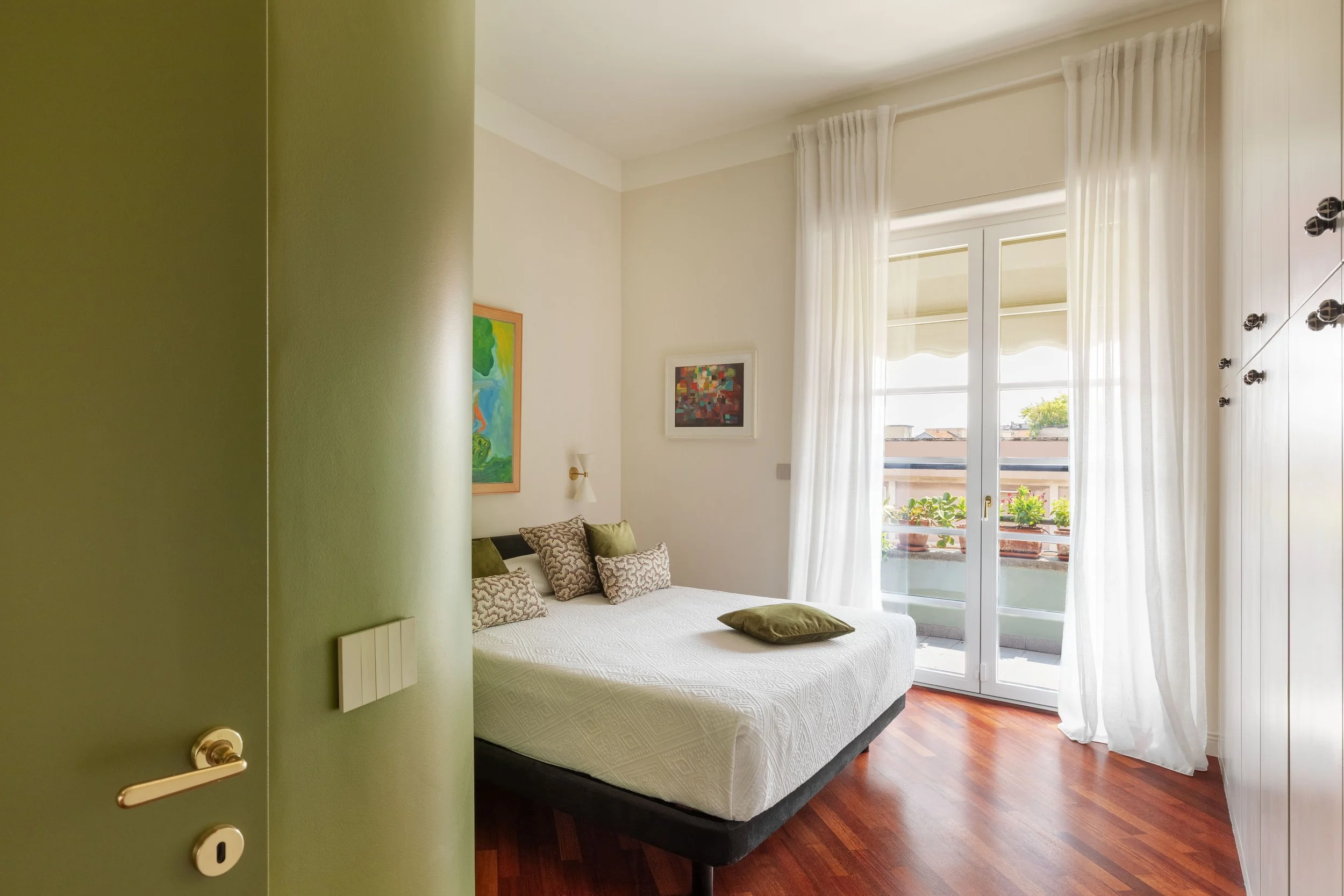 Vista di una camera da letto con letto matrimoniale, cuscini, quadri alle pareti e grande finestra con tende bianche che apre su un balcone con piante e vista sul cielo e altri edifici.