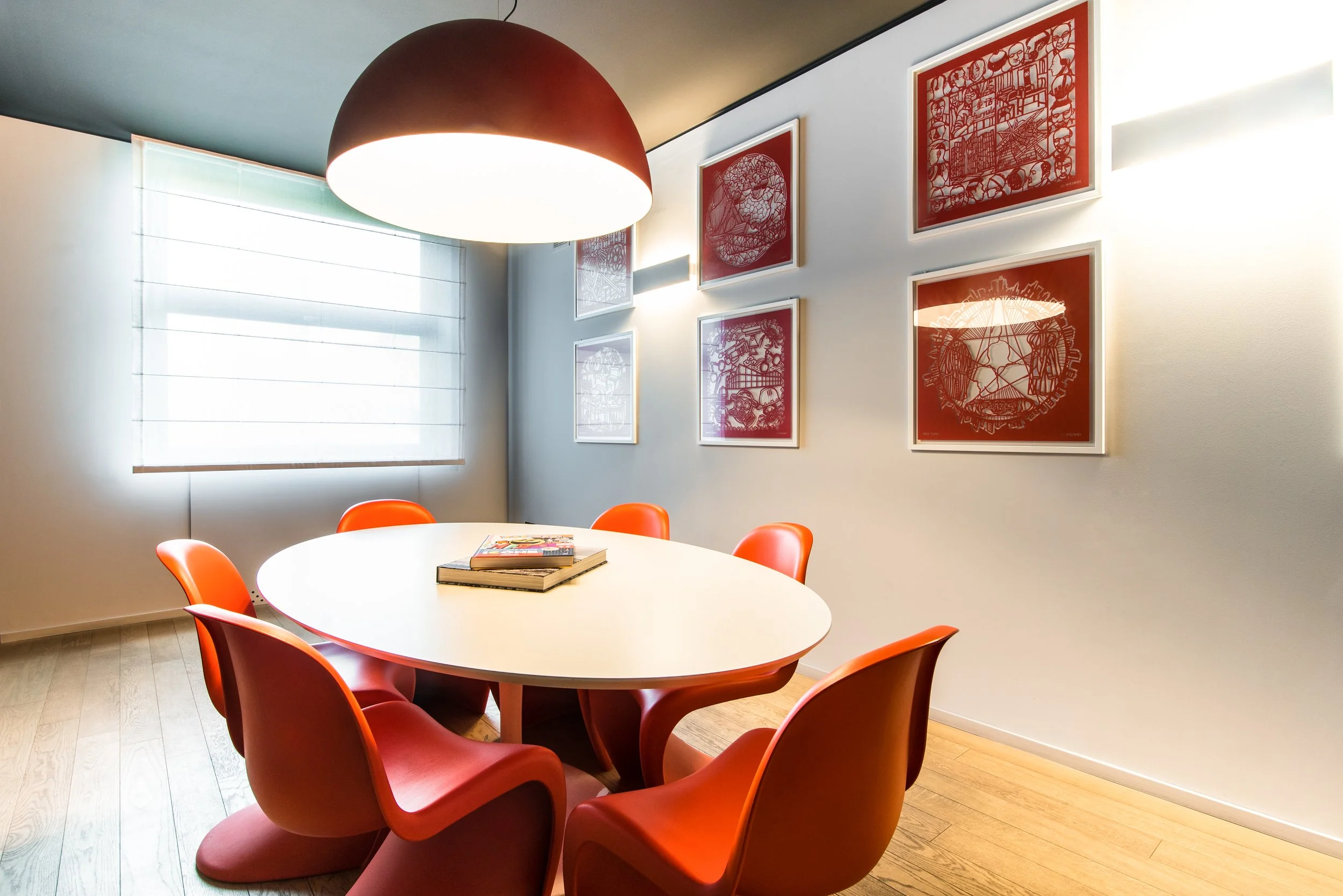 Sala riunioni con tavolo bianco circondato da sei sedie rosse, quadri con sfondo rosso e disegni sulla parete, finestra con tende bianche, grande lampada nera sopra il tavolo, pavimento di legno.