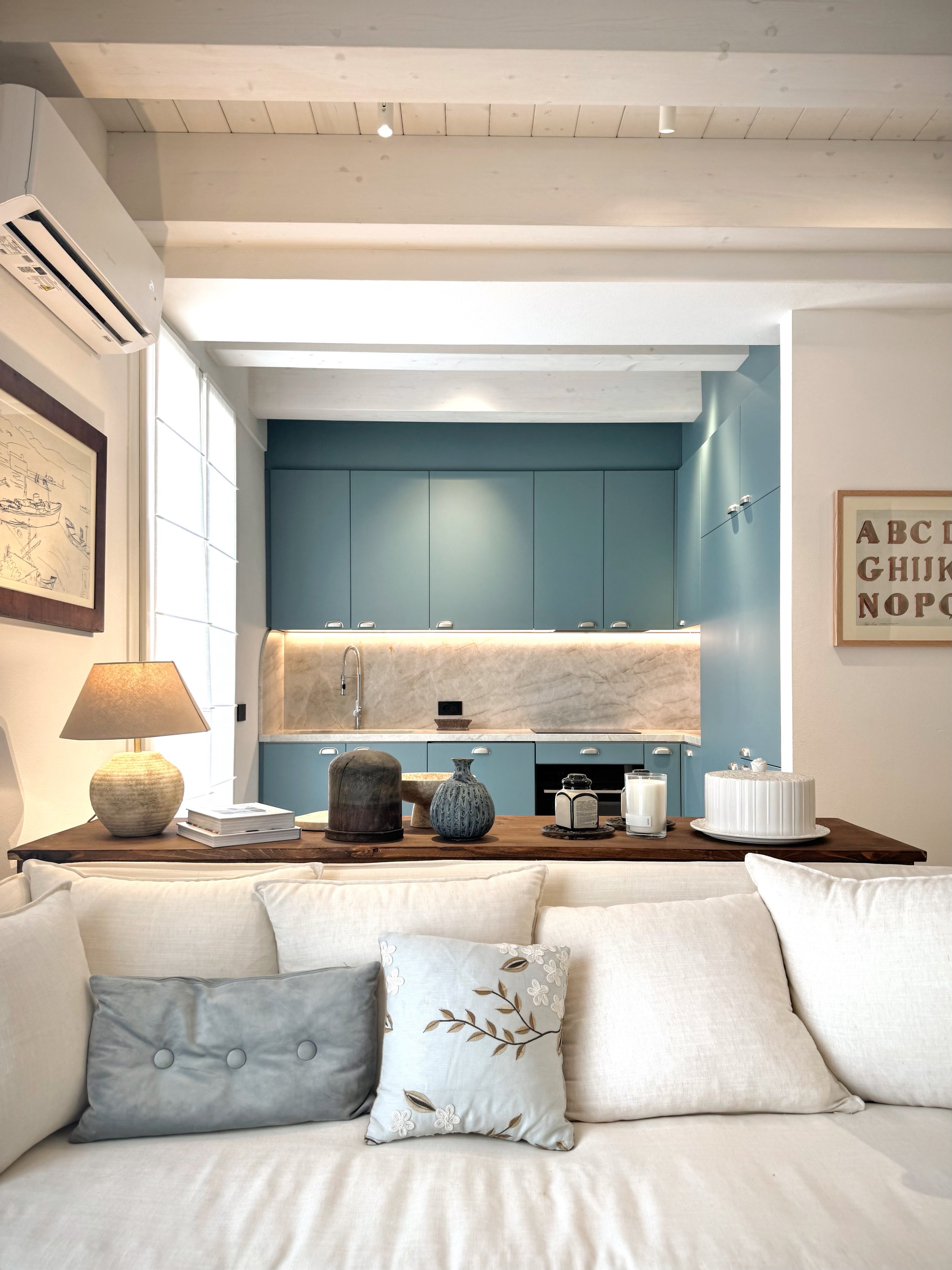 Interno di un soggiorno con divano bianco e cuscini decorativi, tavolo con decorazioni in vasi e candele, cucina moderna sullo sfondo con ante blu e piano in marmo
