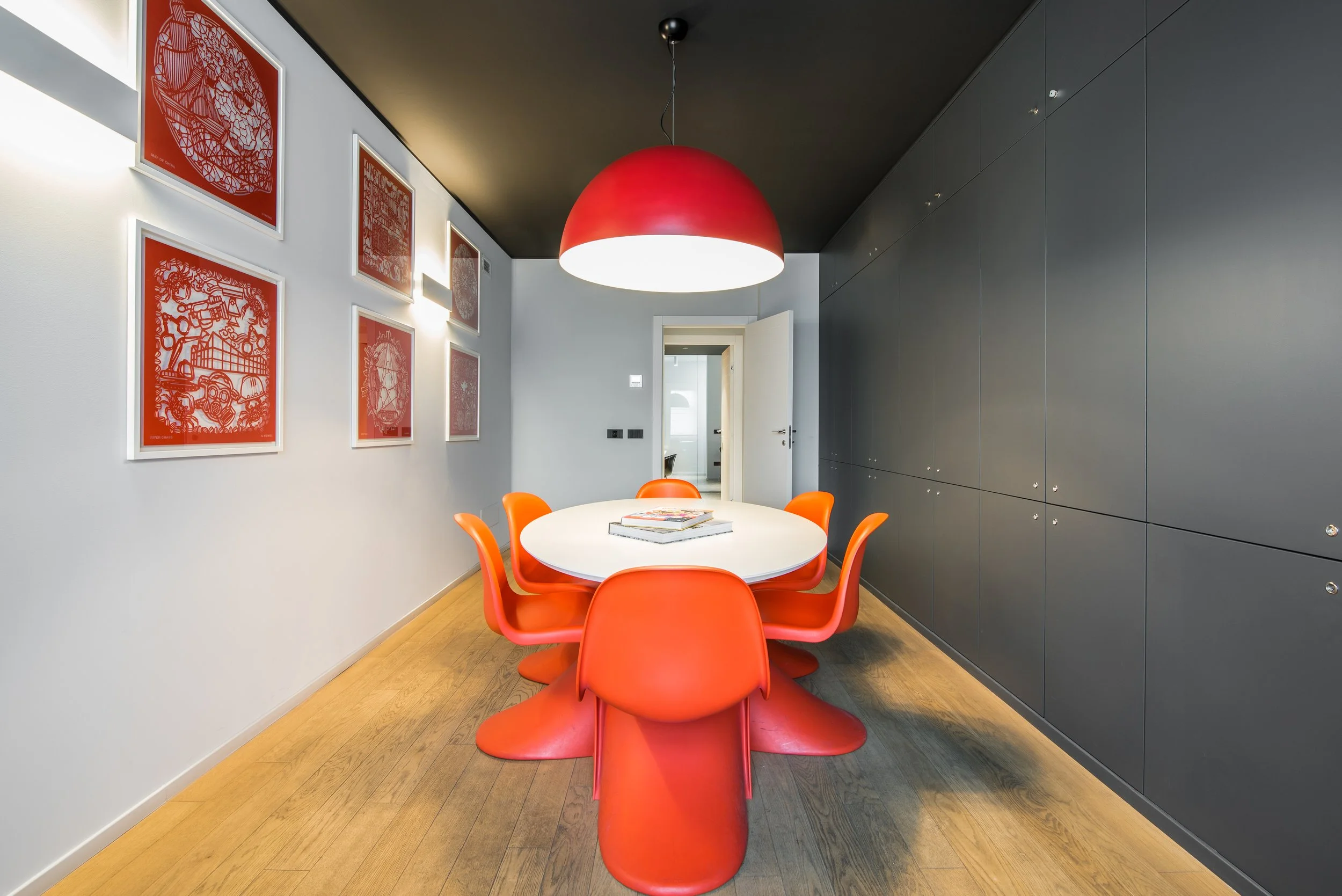 Sala da pranzo moderna con tavolo bianco rotondo, sedie arancioni, grande lampadario rosso, pareti bianche e una parete nera con armadi.