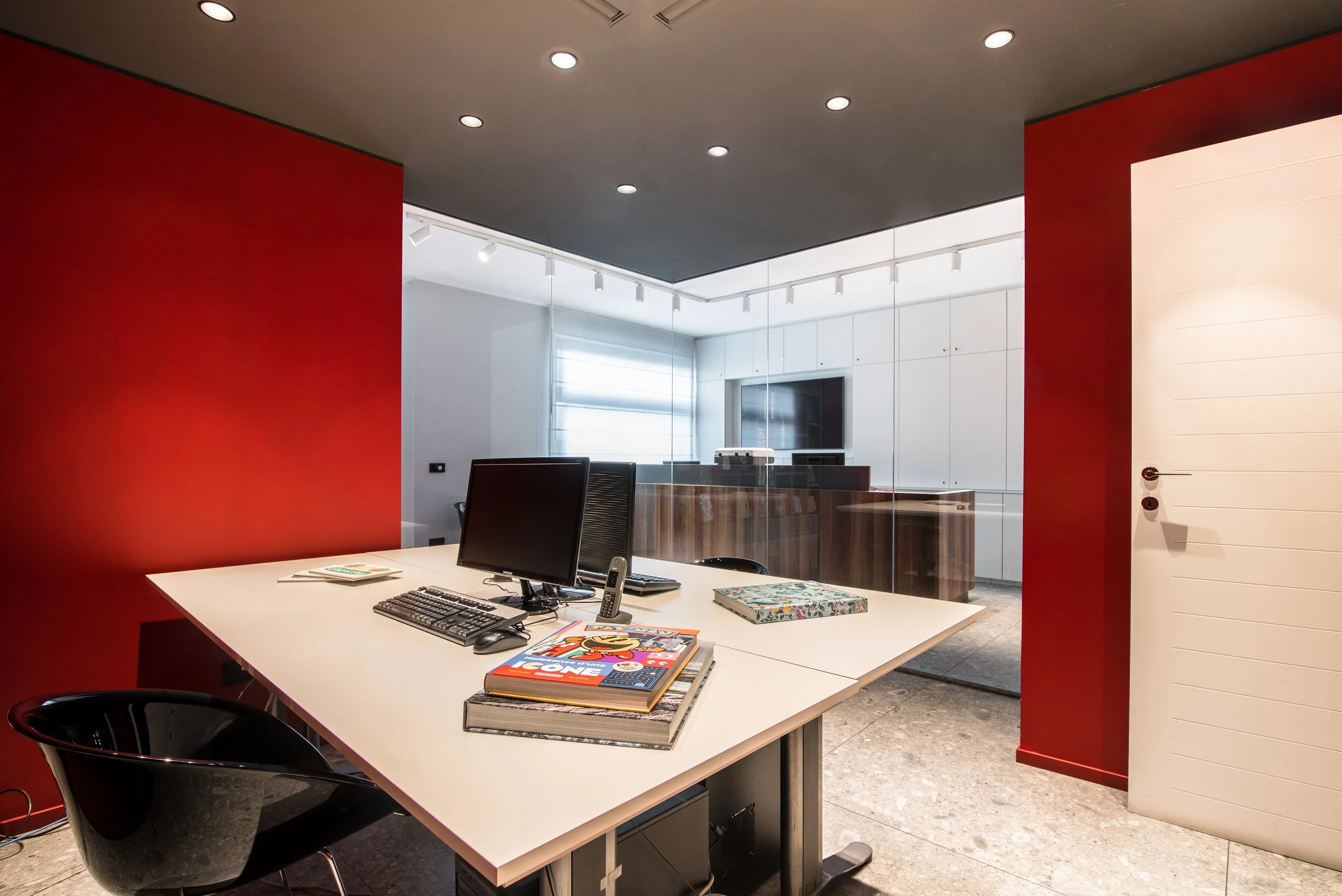 Ufficio moderno con pareti rosse e una zona separata con vetro trasparente, scrivania con computer, tastiera, telefono e riviste.