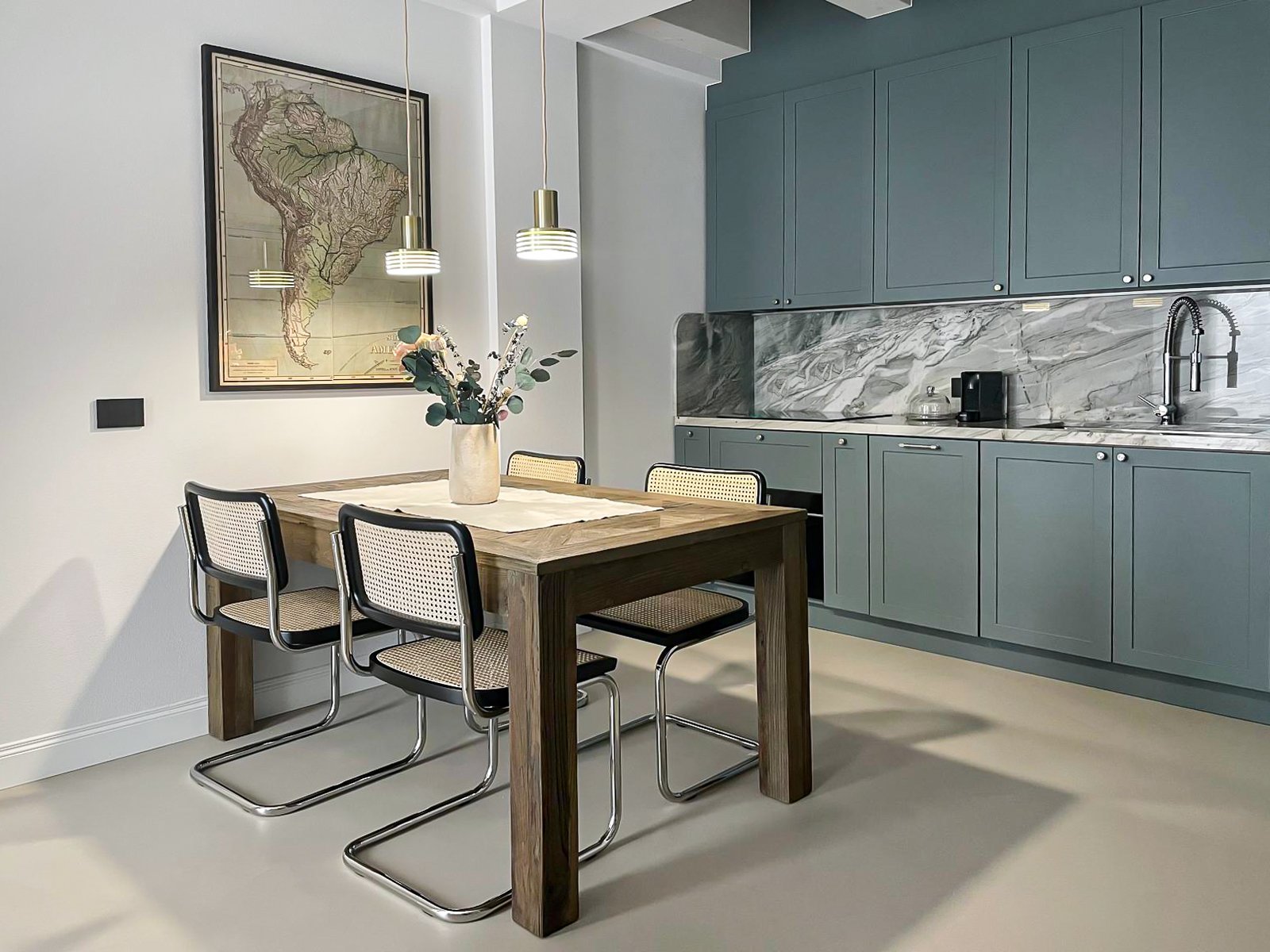 Una cucina moderna con armadietti azzurri, un tavolo in legno con quattro sedie e un vaso con fiori al centro, un quadro con una mappa dell'America del Sud sulla parete e tre lampade a sospensione sopra il tavolo.