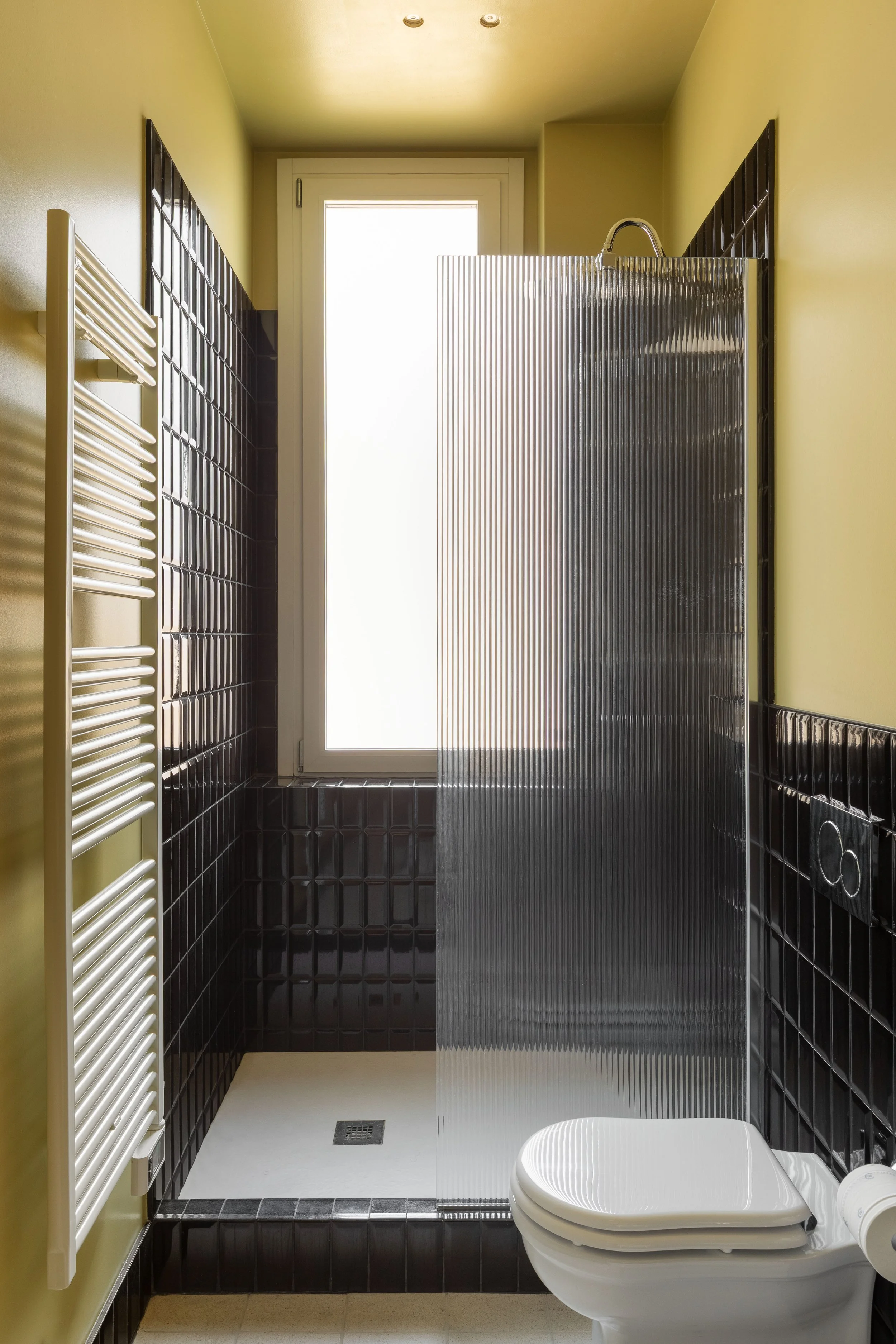 Bagno con pareti rivestite di piastrelle nere, doccia con parete in vetro rigato, WC con coperchio aperto, termosifone bianco e finestra sul soffitto.