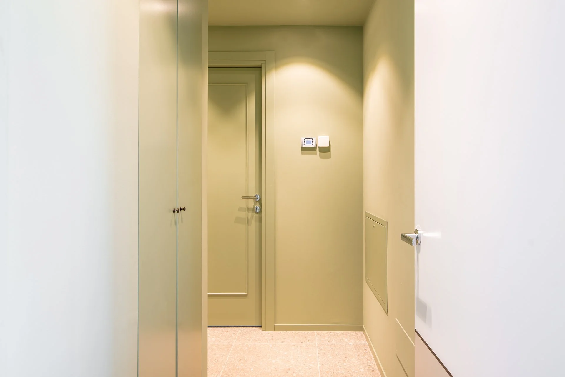 Configurazione di un ingresso o corridoio con pareti verdi chiaro, armadi o armadietti laterali, porta chiusa e numerosi pannelli di controllo o interruttori sulla parete di sfondo.