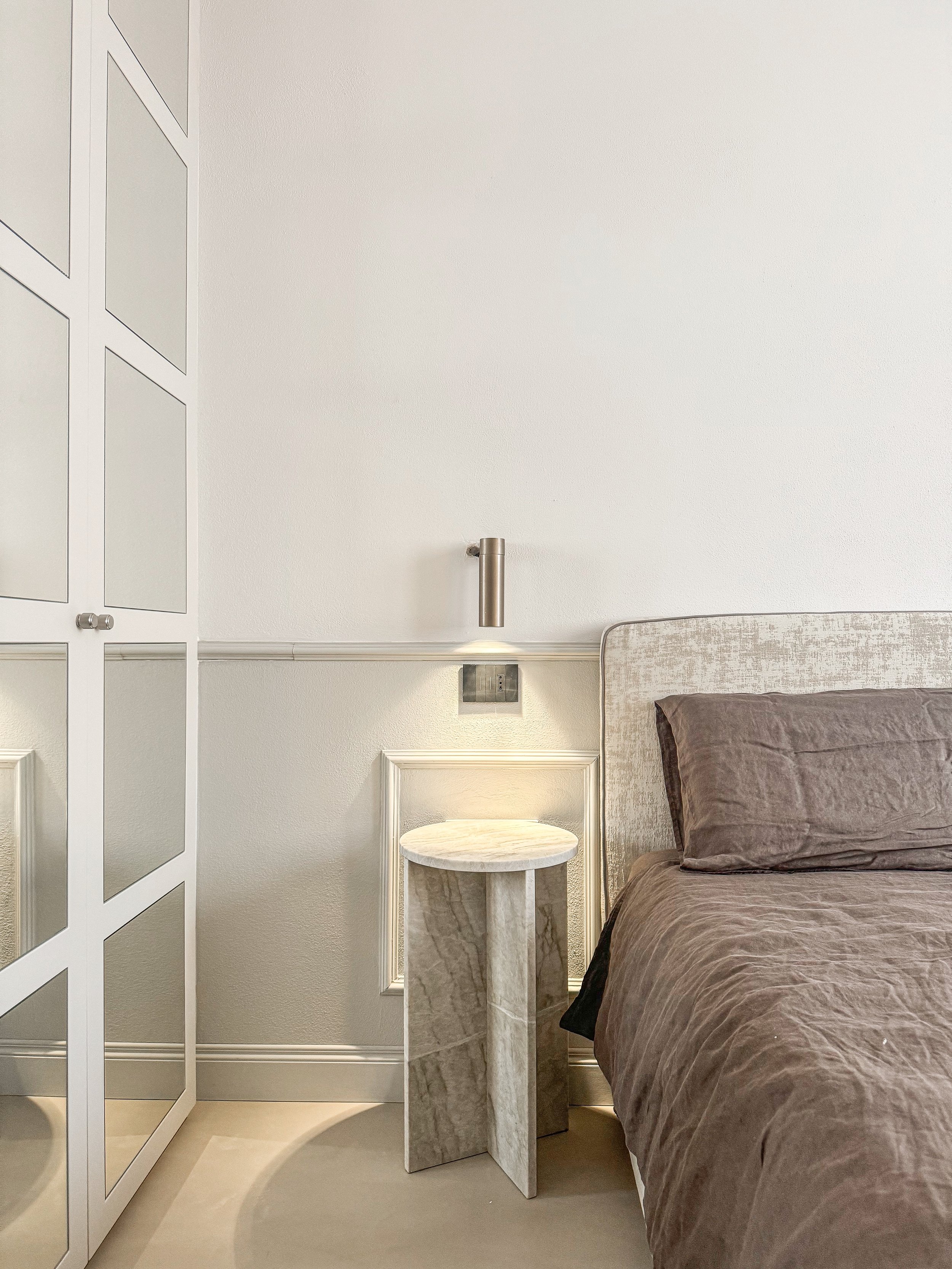 Letto matrimoniale con lenzuola marroni, testiera imbottita, piccola consolle in marmo a fianco, pareti bianche con dettagli architettonici e lampada a parete