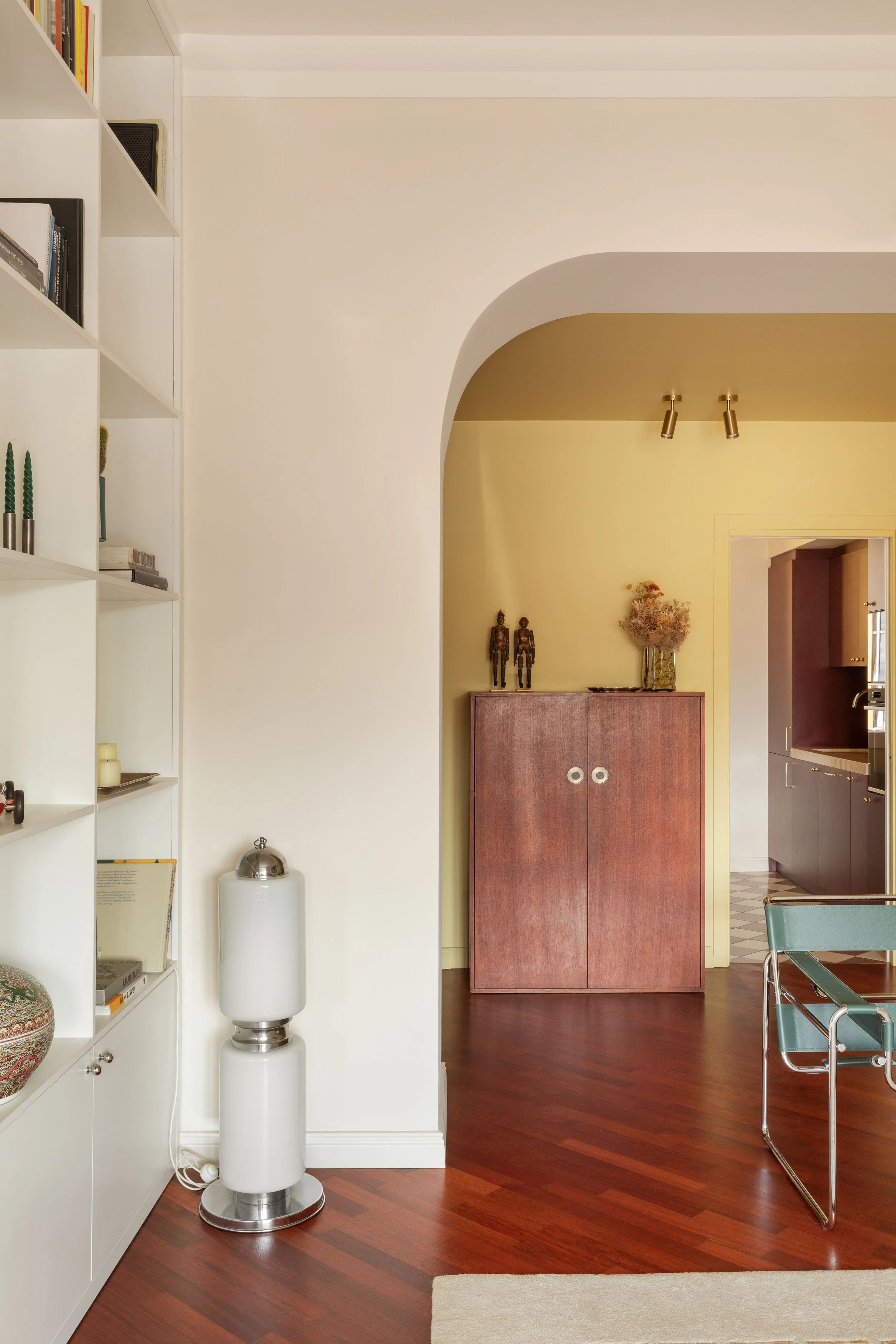 Interno di una stanza moderna con pareti beige e arancio, libreria bianca a sinistra, mobile in legno scuro, specchio, decorazioni, sedie e un'area con cucina visibile in fondo.