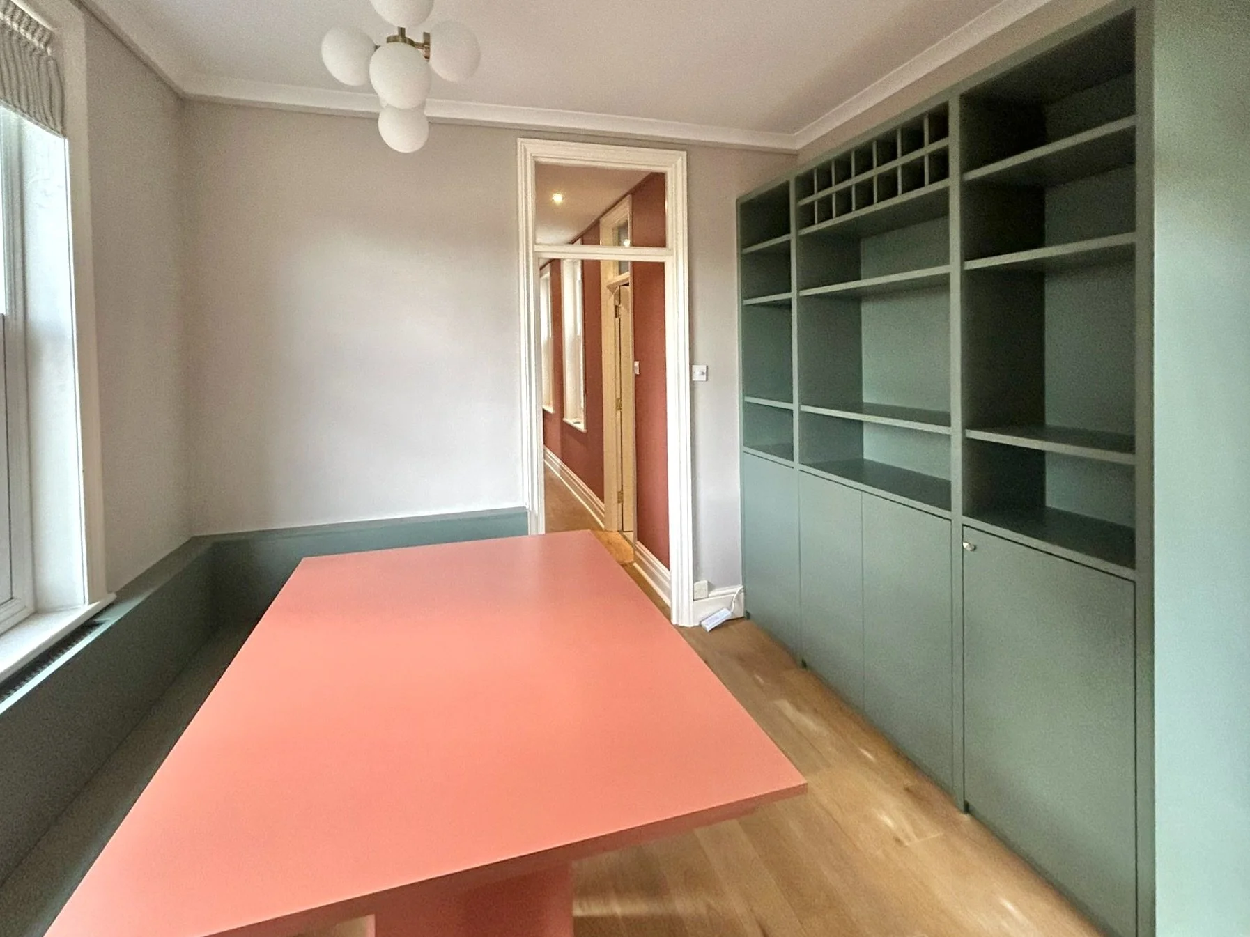 Stanza con pareti grigie, grande finestra, tavolo rosa e grande scaffale verde, pavimenti in legno, ingresso visibile attraverso una porta.