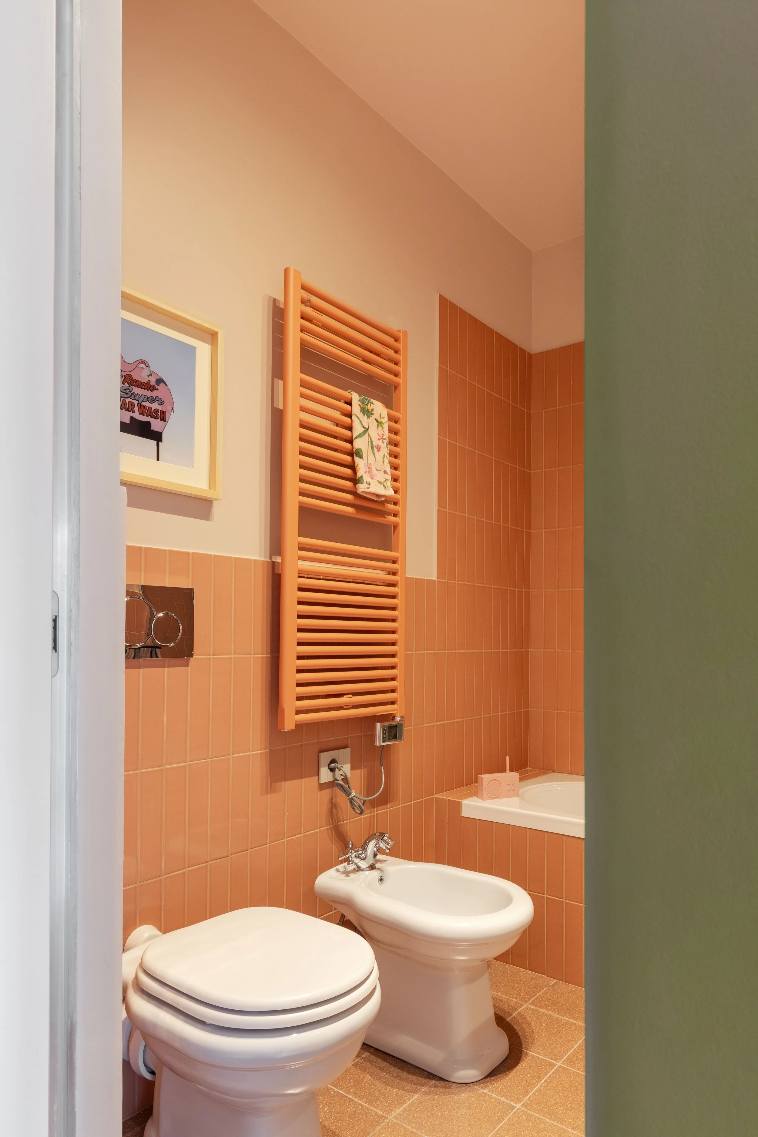 Bagno con piastrelle arancioni, water, bidet, asciugamano su scaldasalviette, quadro appeso, presa elettrica, porta di ingresso, e dettagli del bagno come specchio, vasca e altri accessori.