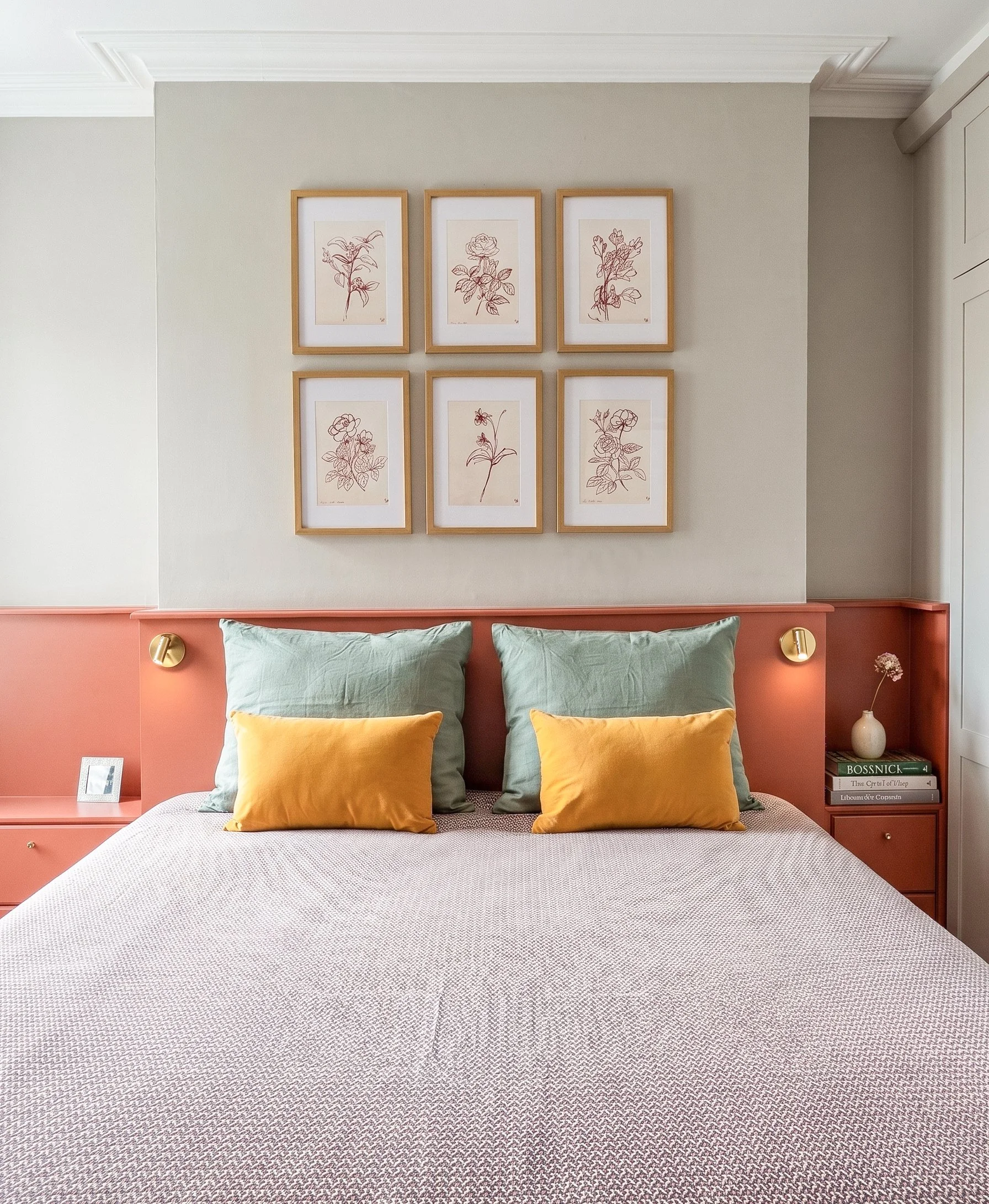 Letto matrimoniale con testiera rosa, quattro cuscini verdi con due cuscini gialli, lampada a muro in ottone ai lati del letto, spalle di un divano o poltrona e una pila di libri sul comodino di destra.