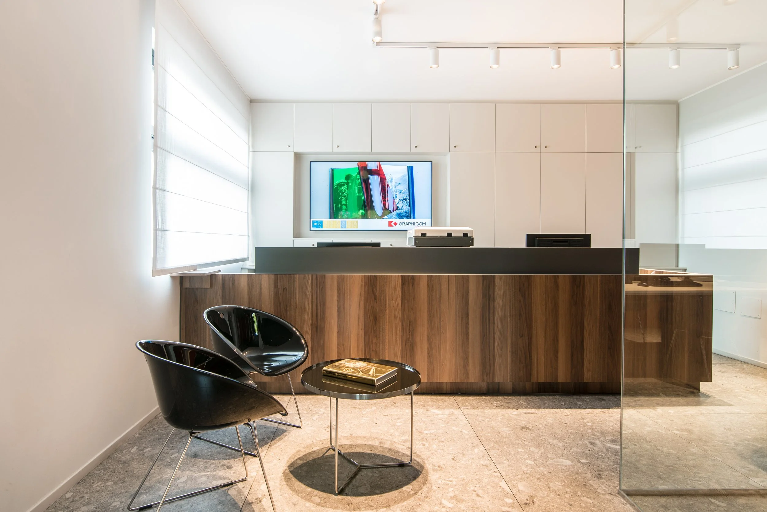 Reception area con bancone in legno scuro, due sedie nere con tavolino centrale e schermo televisivo sulla parete bianca con finestra a destra.