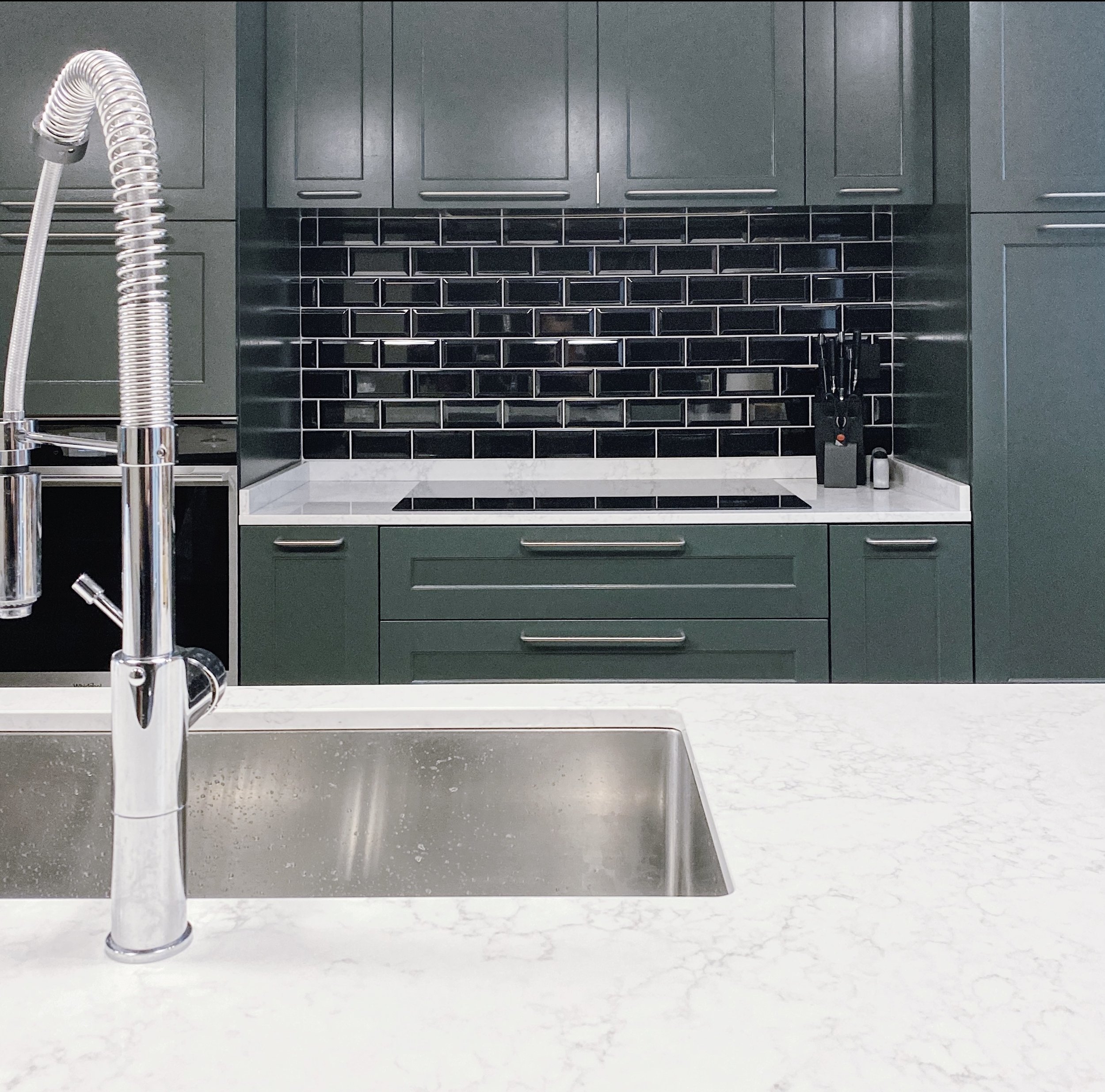 Vista di una cucina moderna con lavabo a destra, ante verdi scure e sfondo con mattonelle nere.