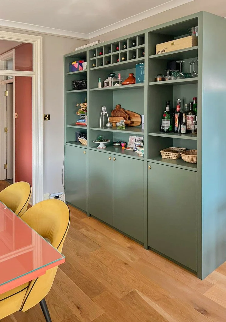 Scaffale verde con bottiglie, bicchieri, decorazioni e oggetti vari in una zona living, con parete bianca e porta a sinistra