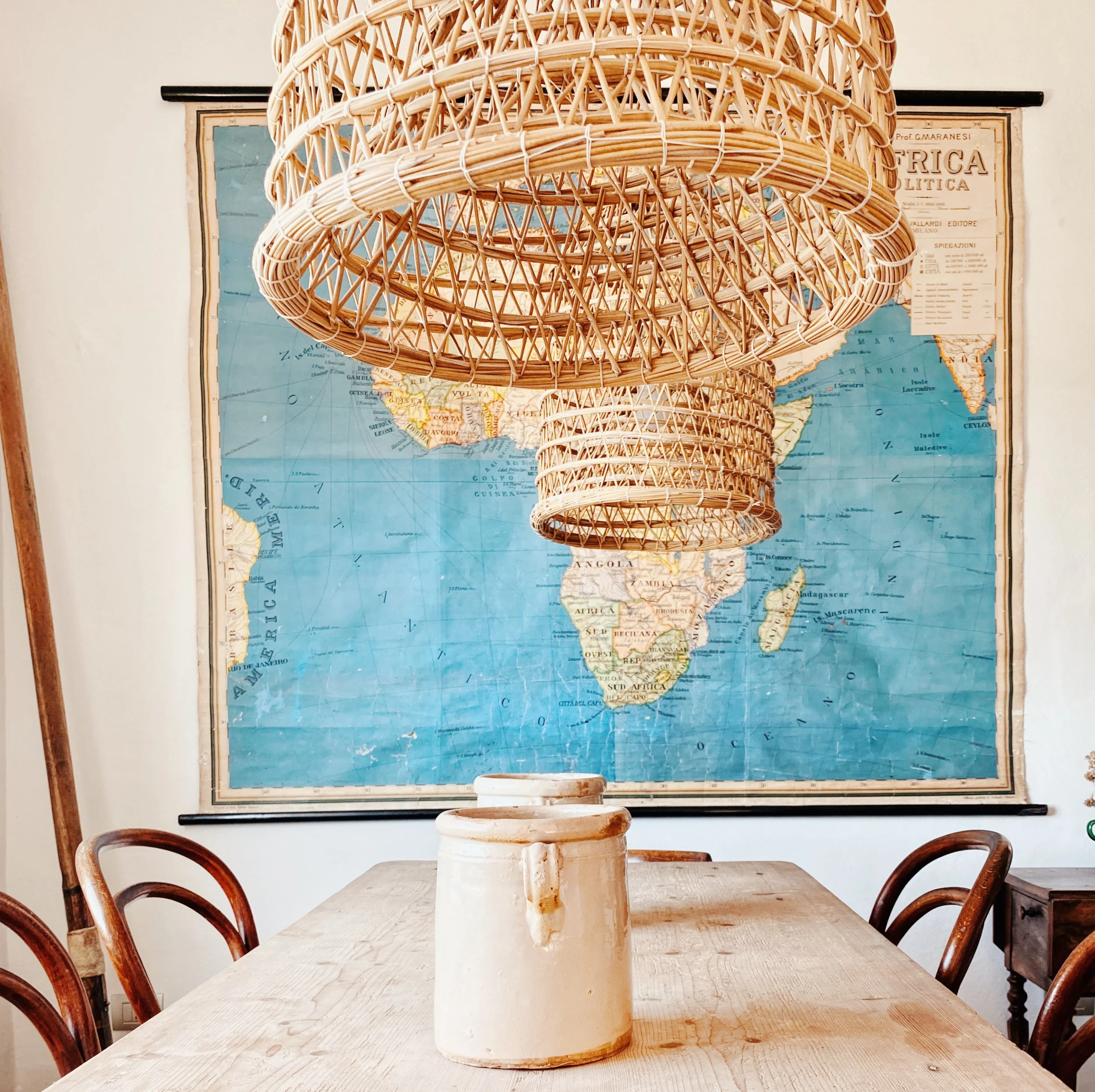 Tavolo di legno con un vaso in ceramica bianca, fondo una mappa del mondo e un lampadario di vimini appeso sopra.