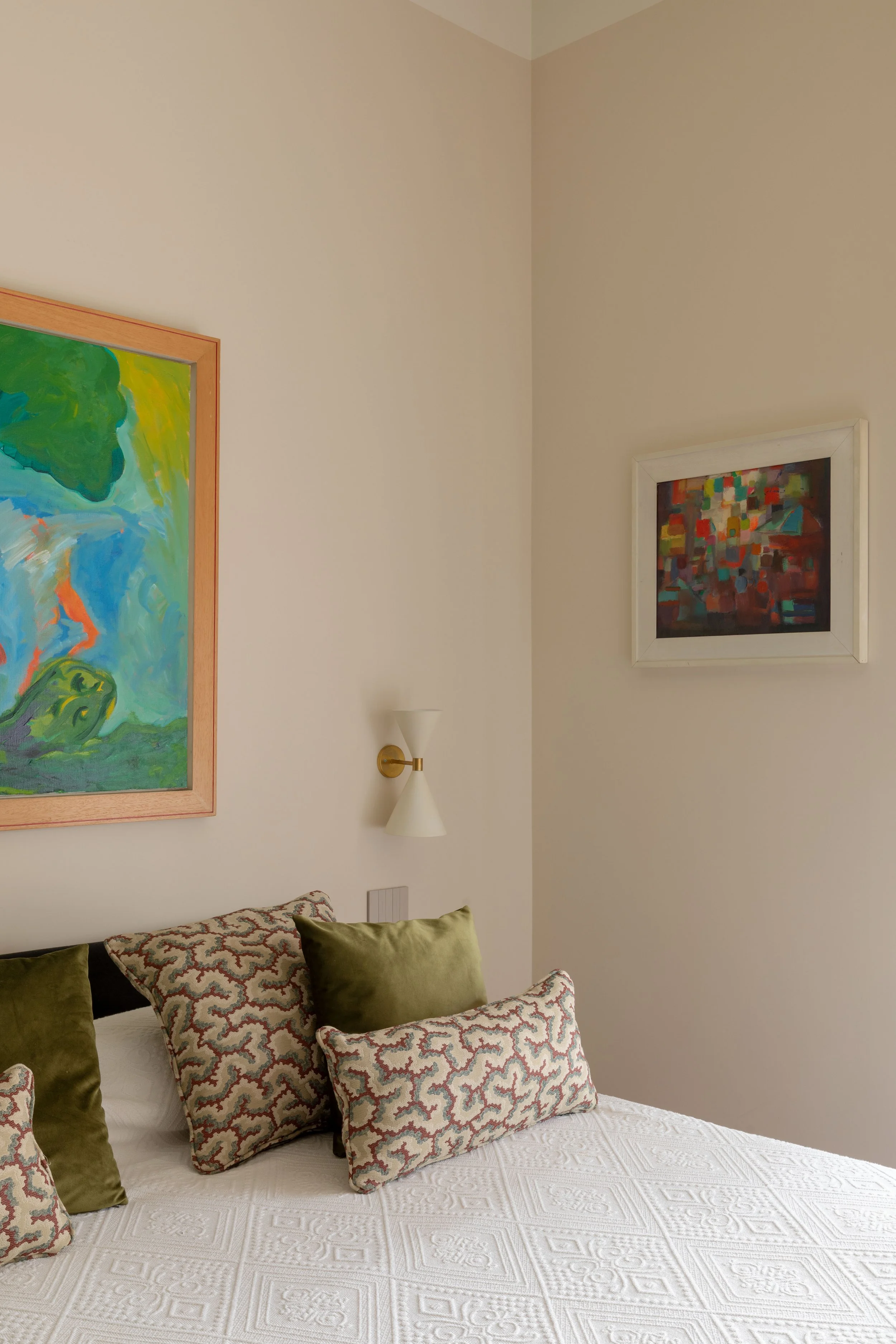 Vista di una stanza con pareti color beige, decorata con quadri astratti, alcuni con cornice di legno chiaro e altri bianchi, un lampadario da parete bianco e un letto con cuscini di velluto verde e cuscini decorativi a motivo astratto.
