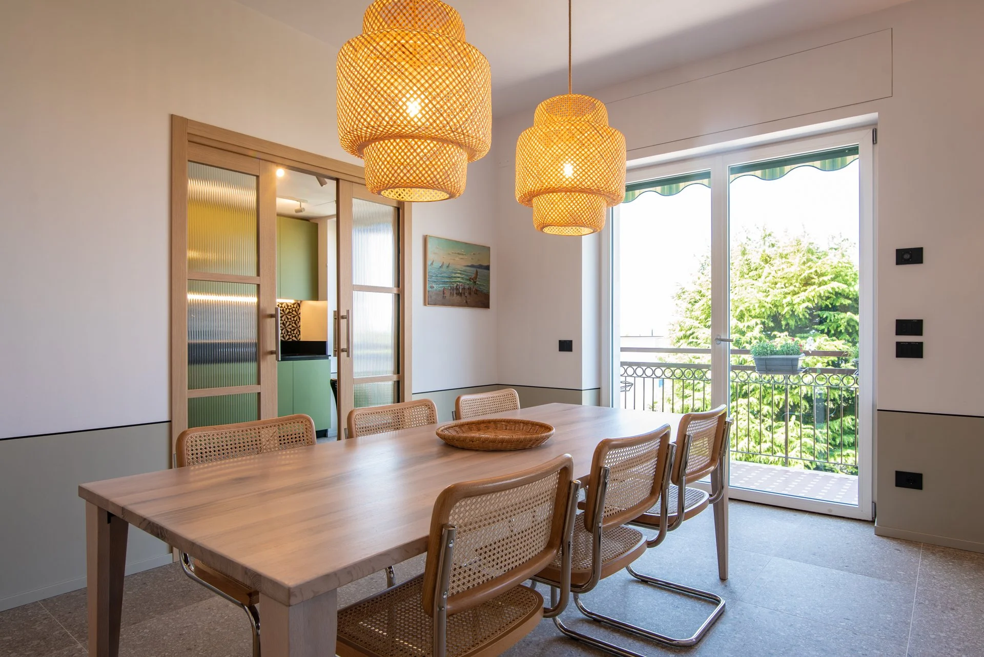 Sala da pranzo con tavolo di legno, sei sedie con seduta in vimini, lampade di vimini appese al soffitto, porta finestra che si affaccia su un balcone con vista su alberi verdi. Illuminato dalla luce naturale.