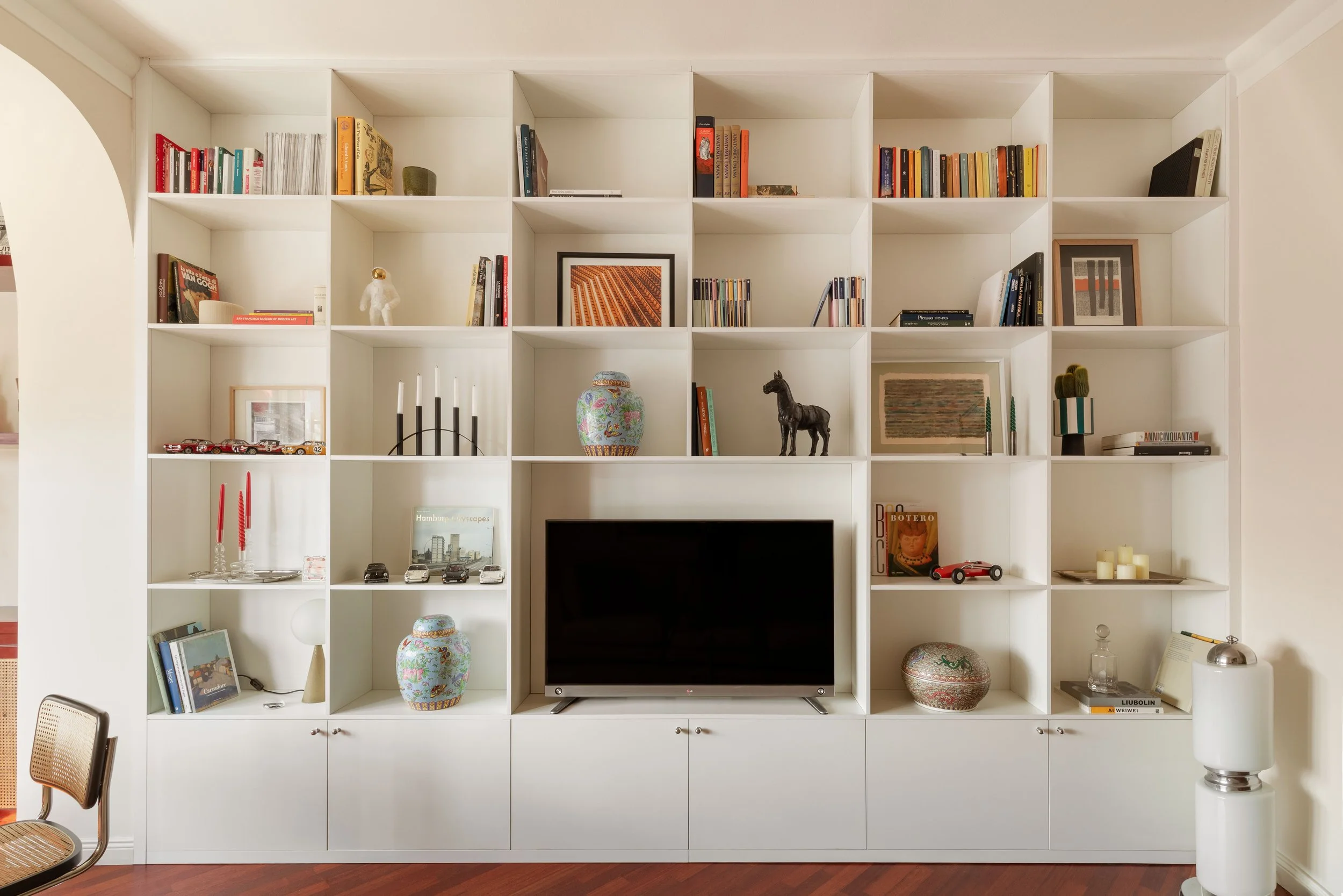 Pensile bianco con scaffali pieni di libri, decorazioni, modellini di automobili e vasi ornamentali. C'è anche un televisore al centro e alcune sedie in primo piano.