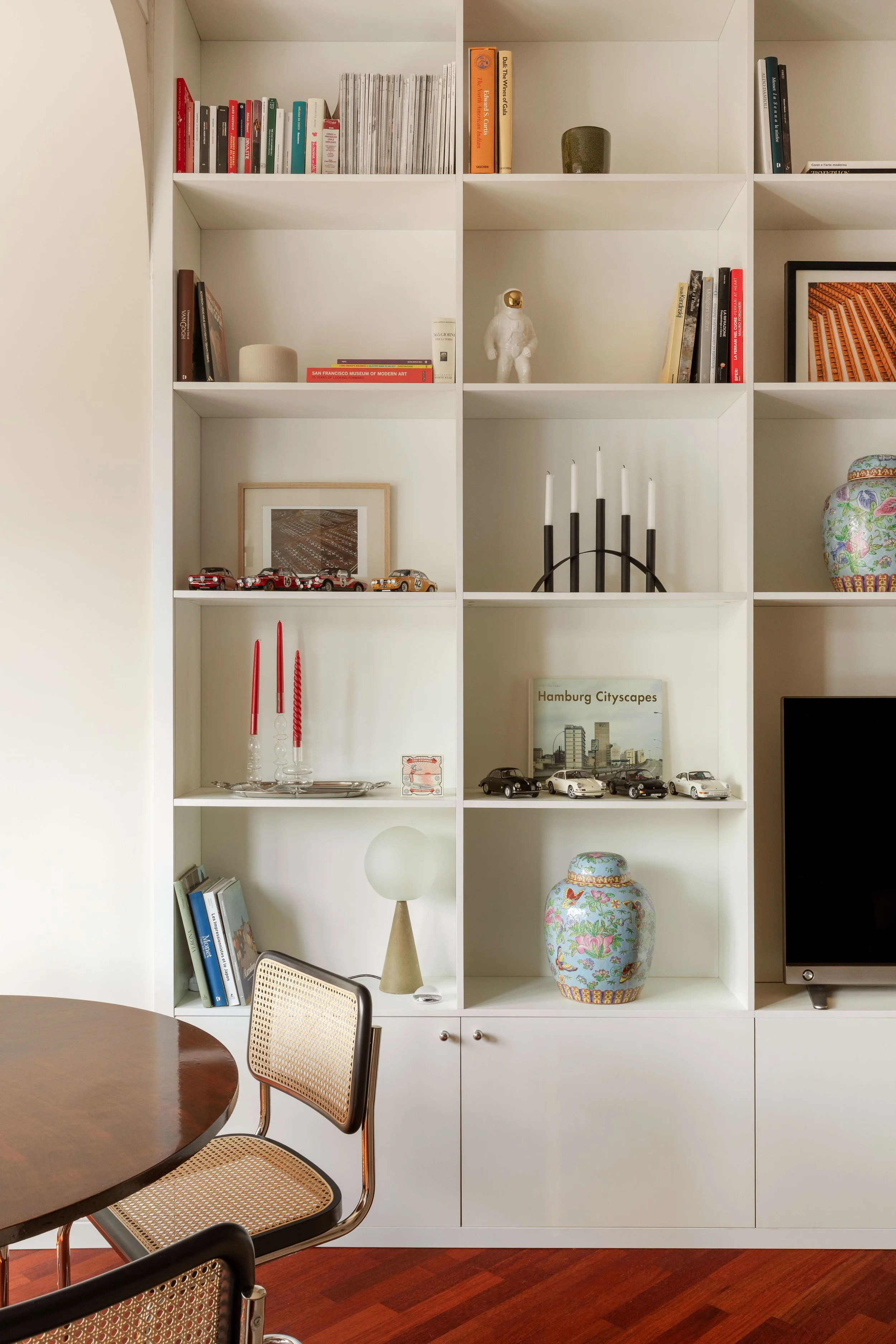 Scaffale bianco con libri, oggetti decorativi e modellini di automobili, tavolo rotondo in legno con sedie in vimini, pavimento in legno scuro.