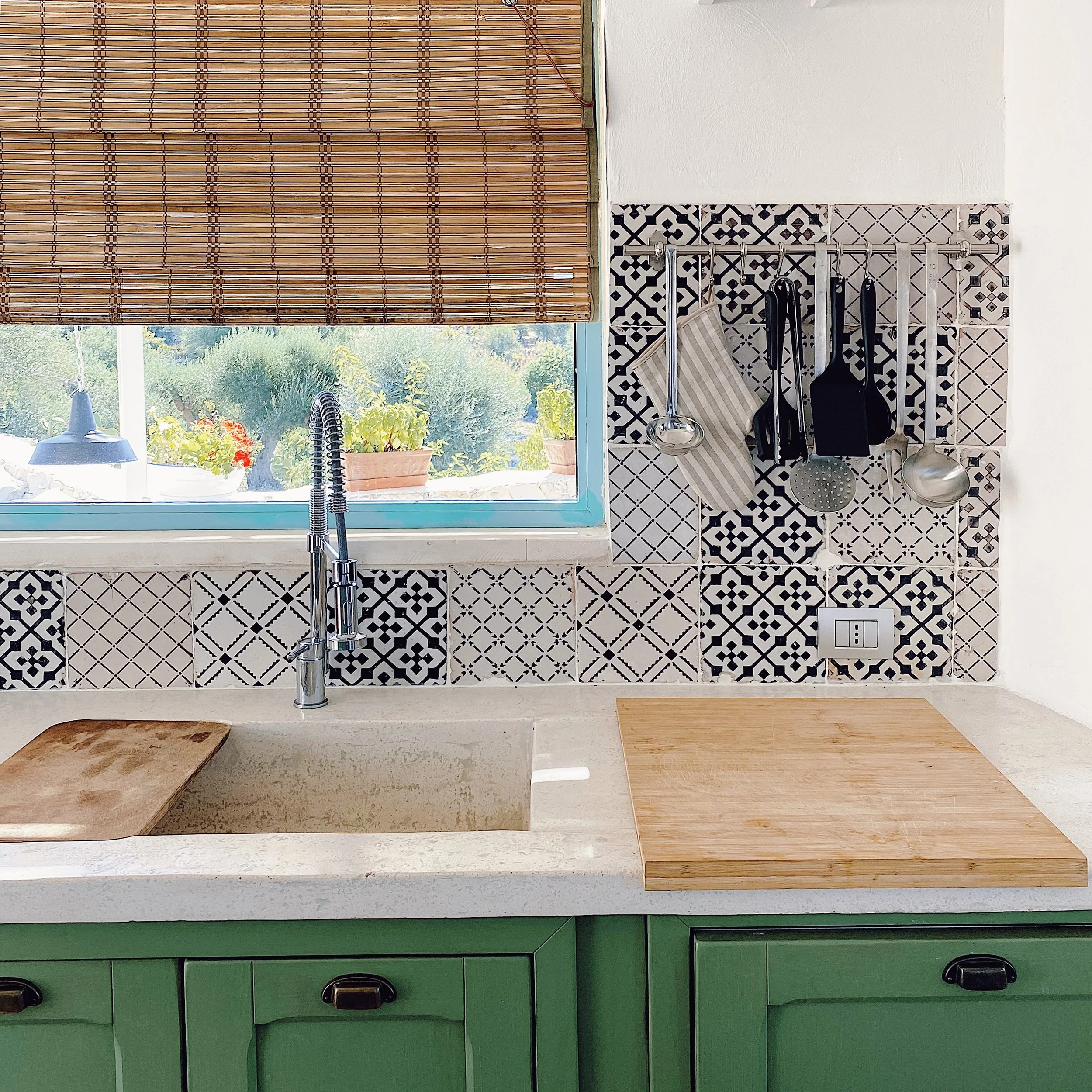 Scorciatoia cucina con lavabo, finestra, tende di bambù, piastrelle decorative nere e bianche, piani di lavoro in pietra, e utensili da cucina appesi al muro.