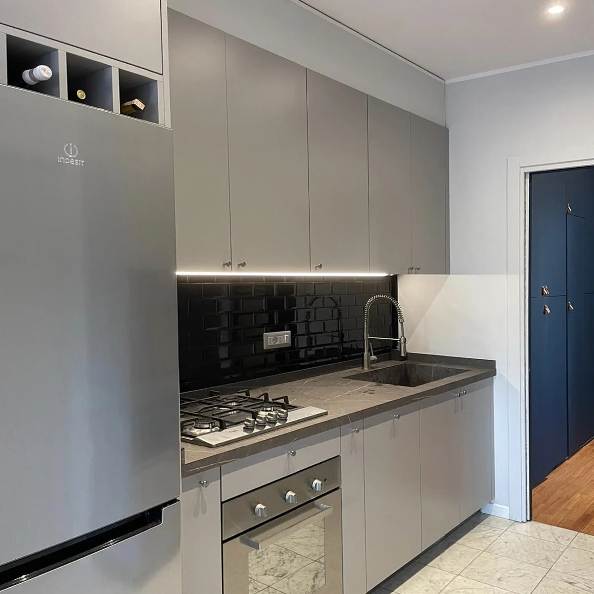 Cucina moderna con frigorifero, piano cottura a quattro fuochi, lavabo con doccia, pensili grigi, parete in piastrelle nere, luci a LED sotto i pensili, porta blu a pannelli, pavimento in piastrelle beige.