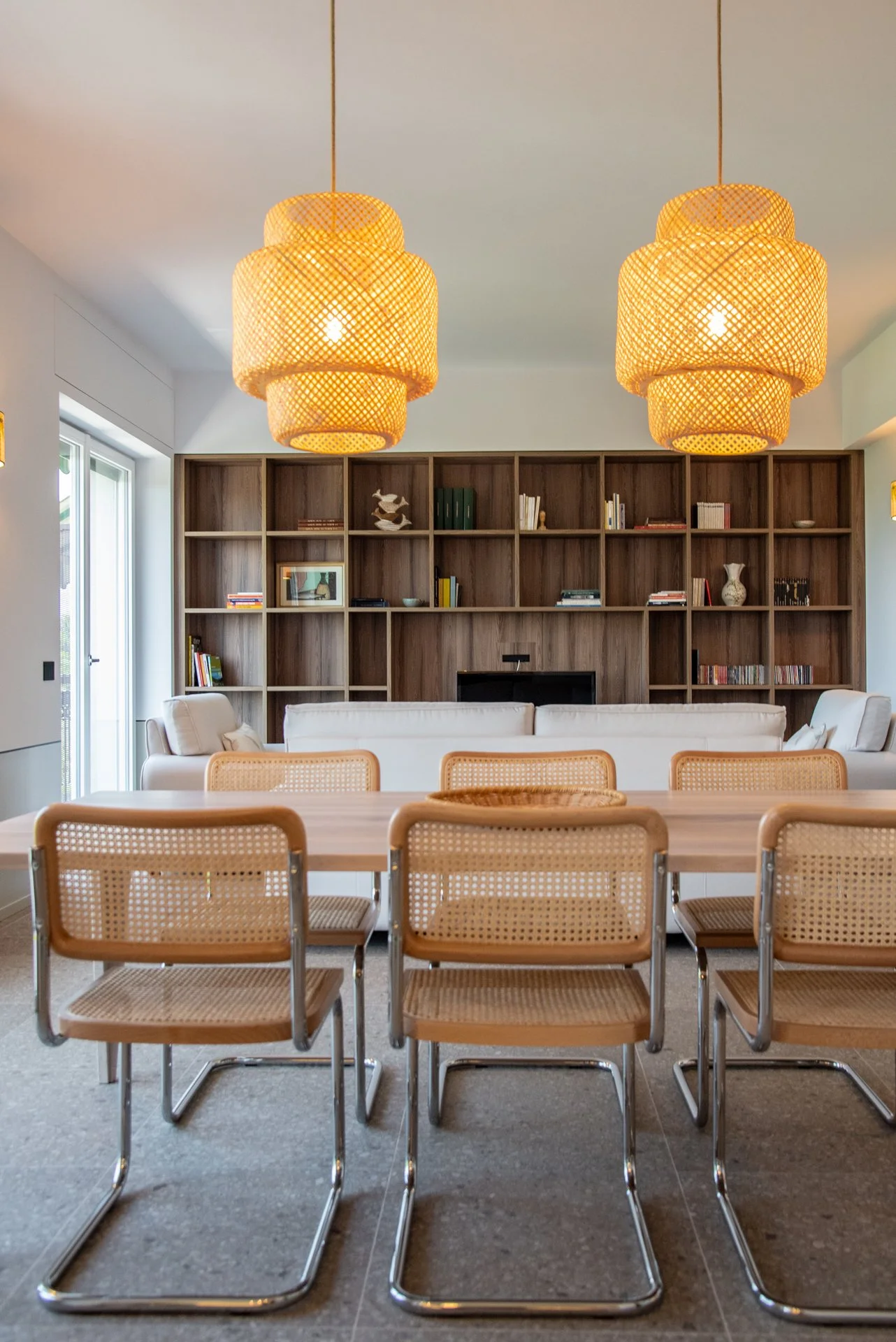Interno di una stanza con tavolo e sedie, libreria in legno sullo sfondo e lampade di vimini suspended.