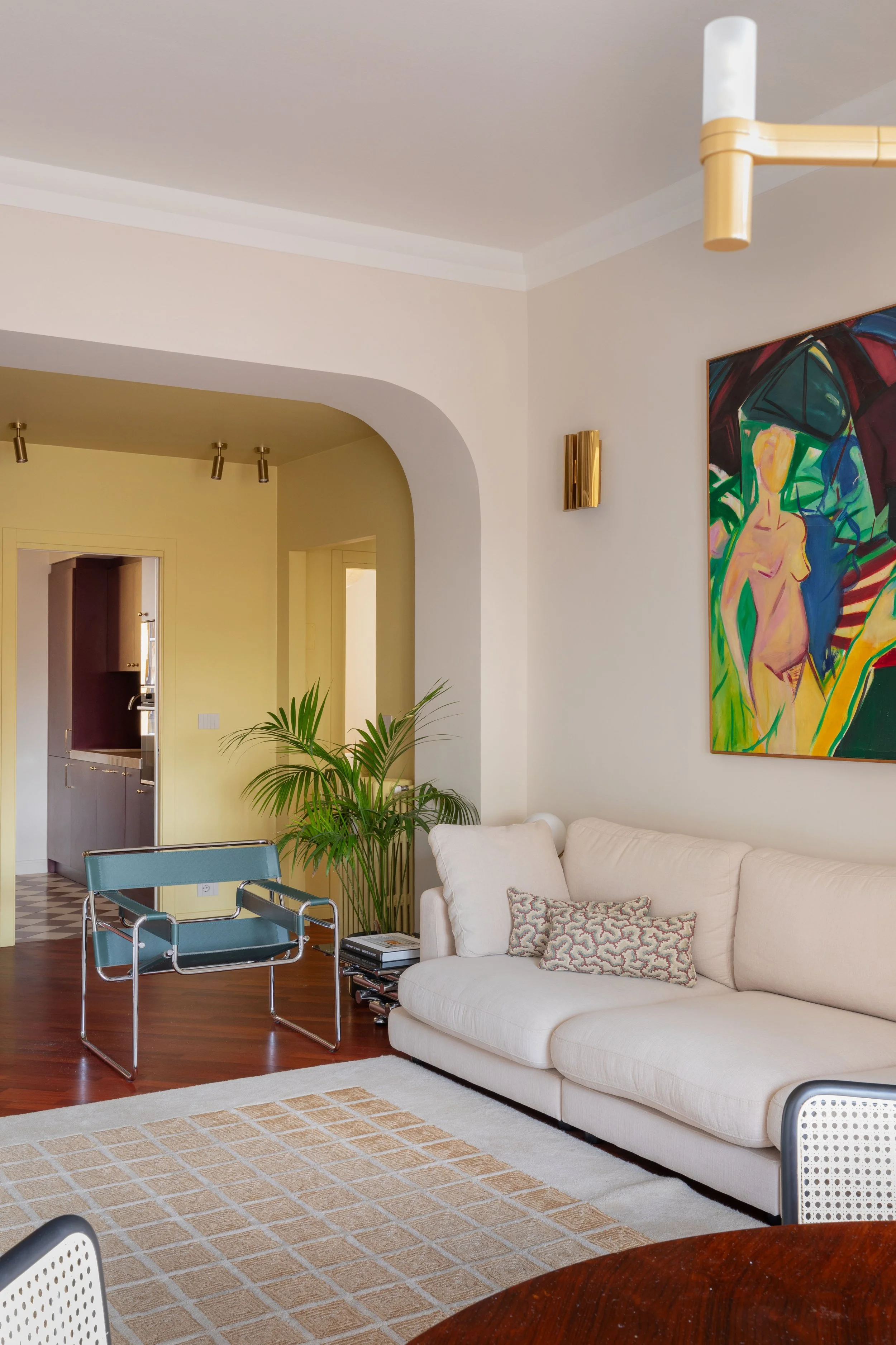 Interno di un soggiorno con divano bianco, una pianta verde, un quadro astratto colorato e un tappeto beige con motivo a reticolo. In sottofondo cucina visibile attraverso un arco.