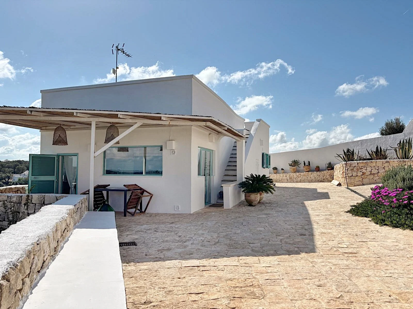 Casa bianca con terrazza, piante e fiori sotto un cielo blu con nuvole