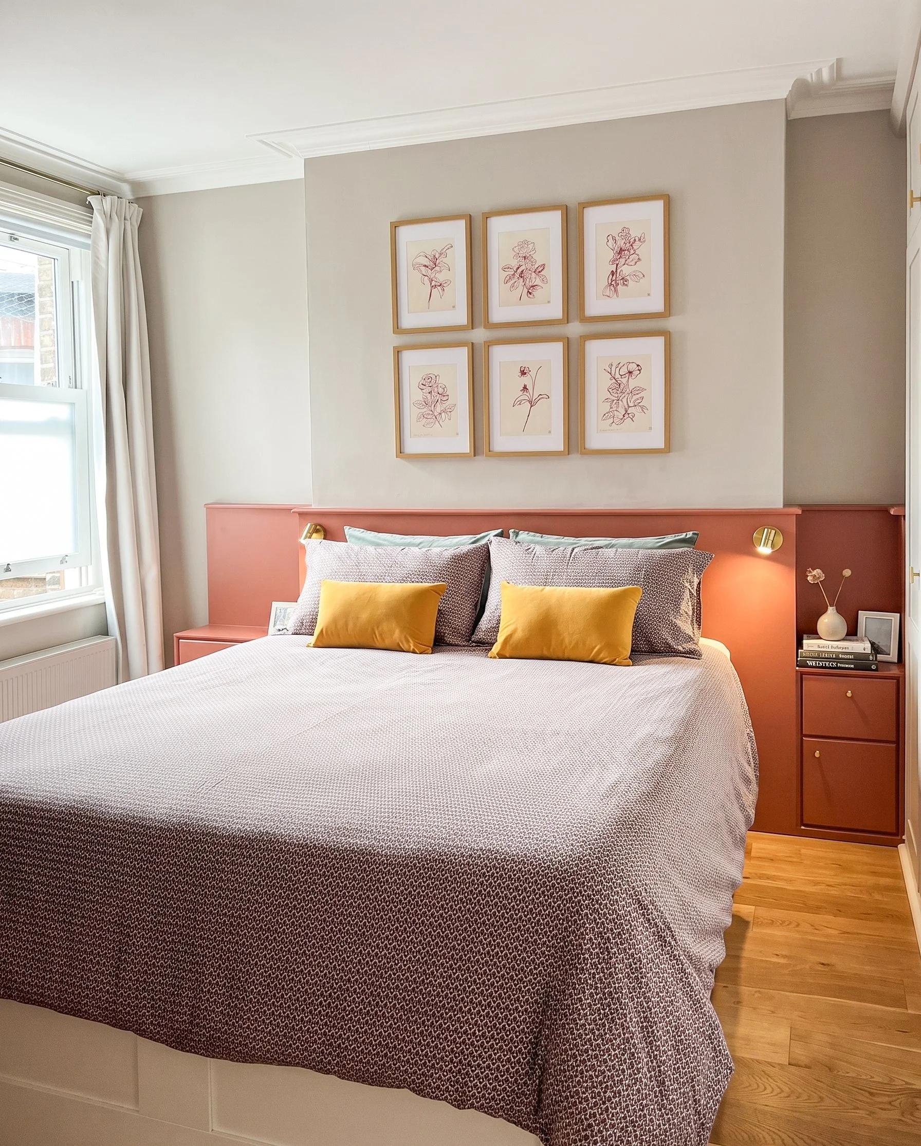 Una camera da letto con un letto matrimoniale con cuscini gialli e bianchi, pareti beige e parete con sei stampe di fiori incorniciate, una finestra con tende bianche, pavimento in parquet, lampada da comodino sul lato destro e una piccola pianta in 