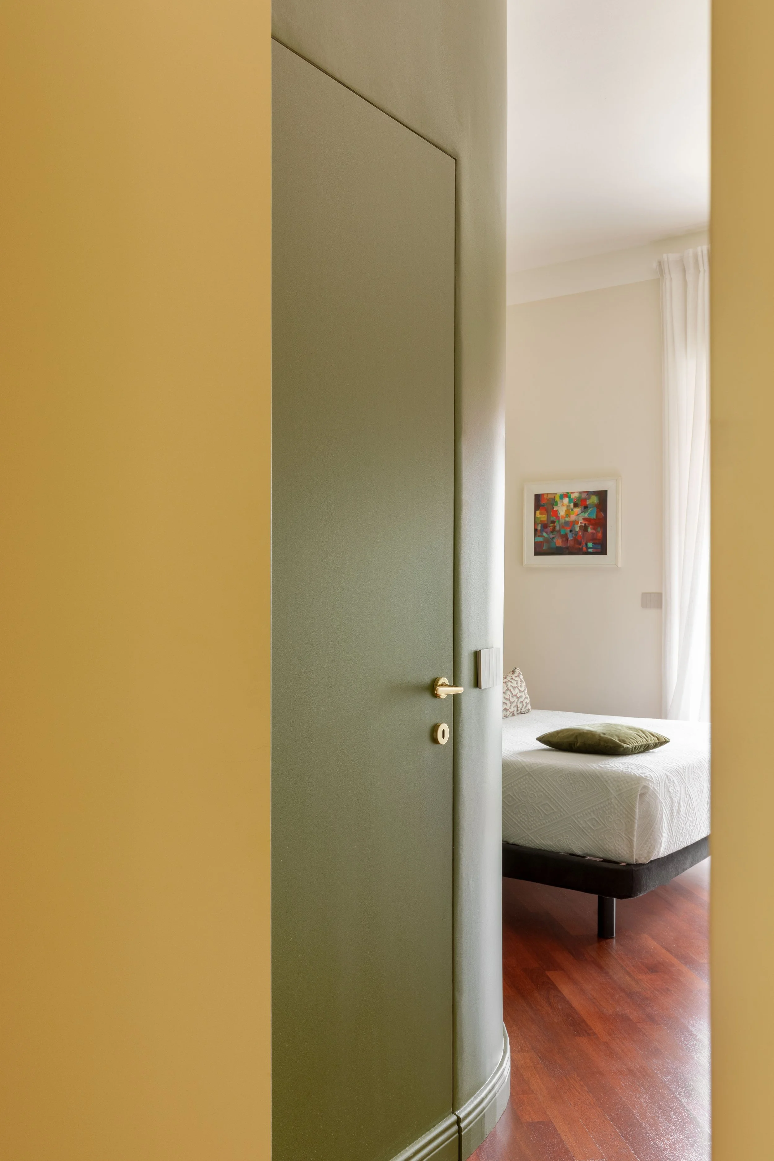 Vista di una stanza con un letto con lenzuola bianche e un cuscino verde. Sul muro una cornice con un dipinto astratto. La stanza è illuminata dalla luce naturale attraverso una finestra con tende bianche.