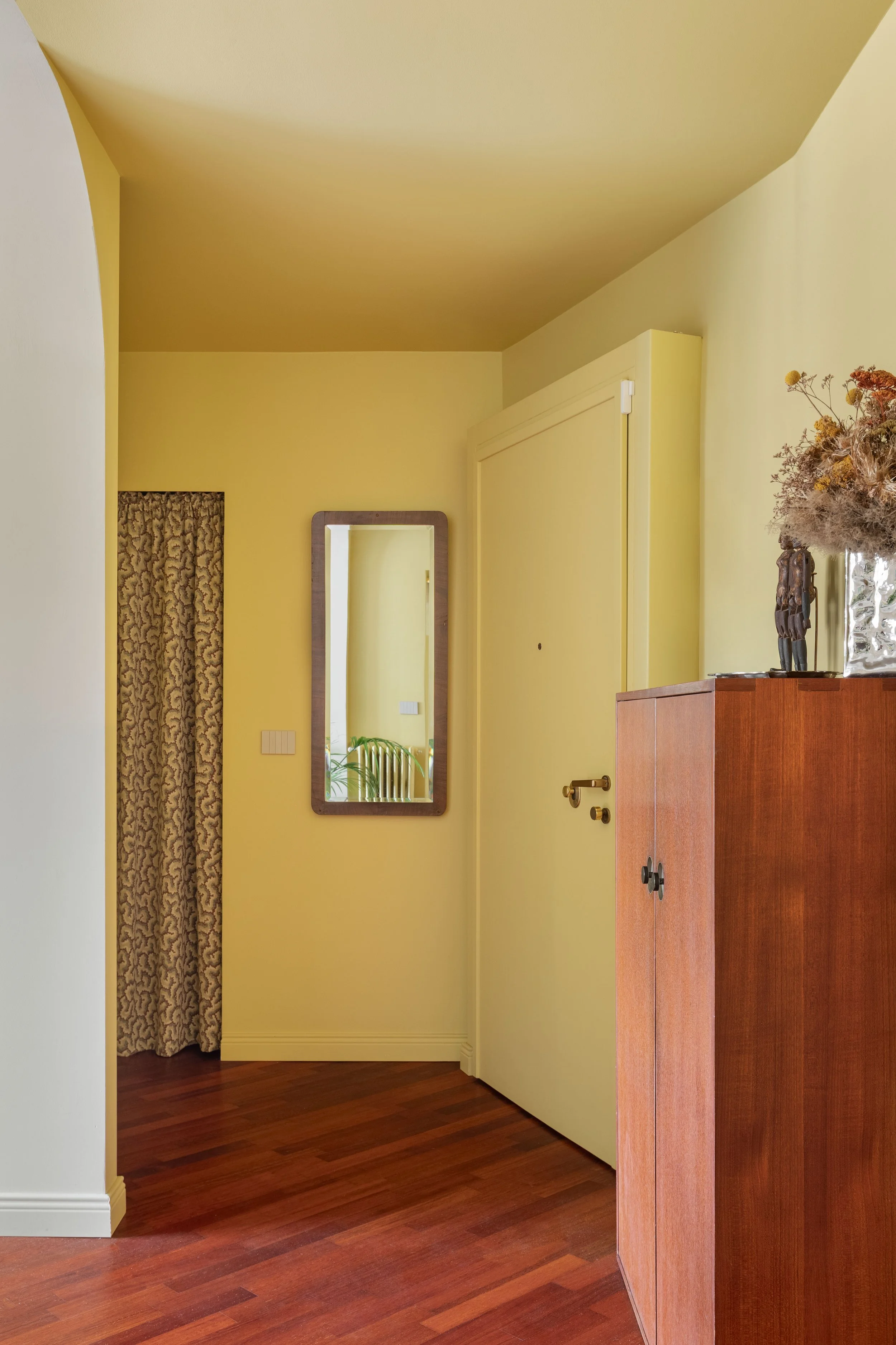 Interno di una stanza con pareti di colore giallo, un mobile di legno, un quadro con cornice di legno sulla parete e una grande pianta con fiori secchi su un tavolo.