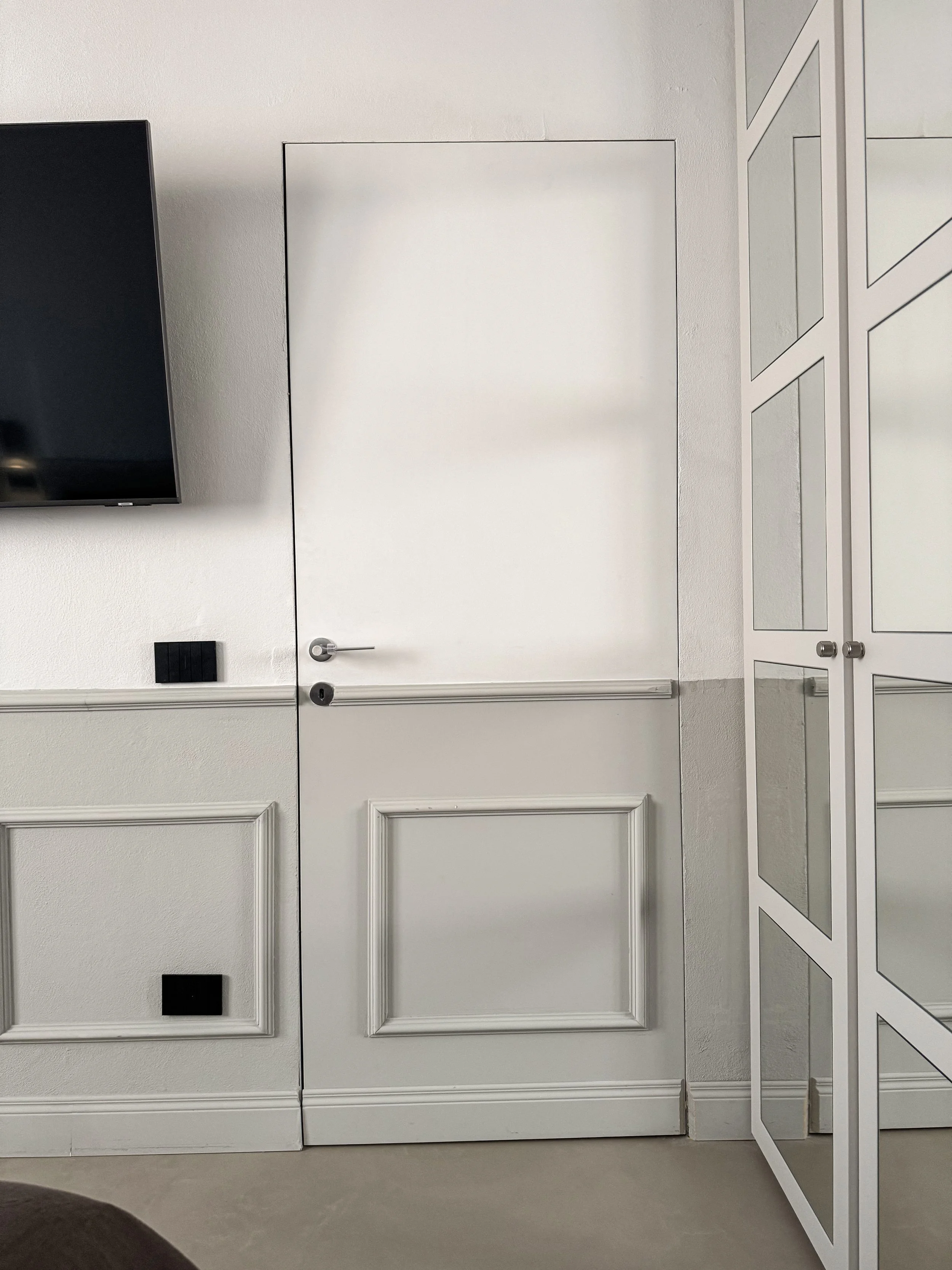 Porta bianca con maniglia in acciaio, parte di una parete bianca con pannelli decorativi, un armadio bianco con ante di vetro, e una televisione a schermo nero montata sulla parete.