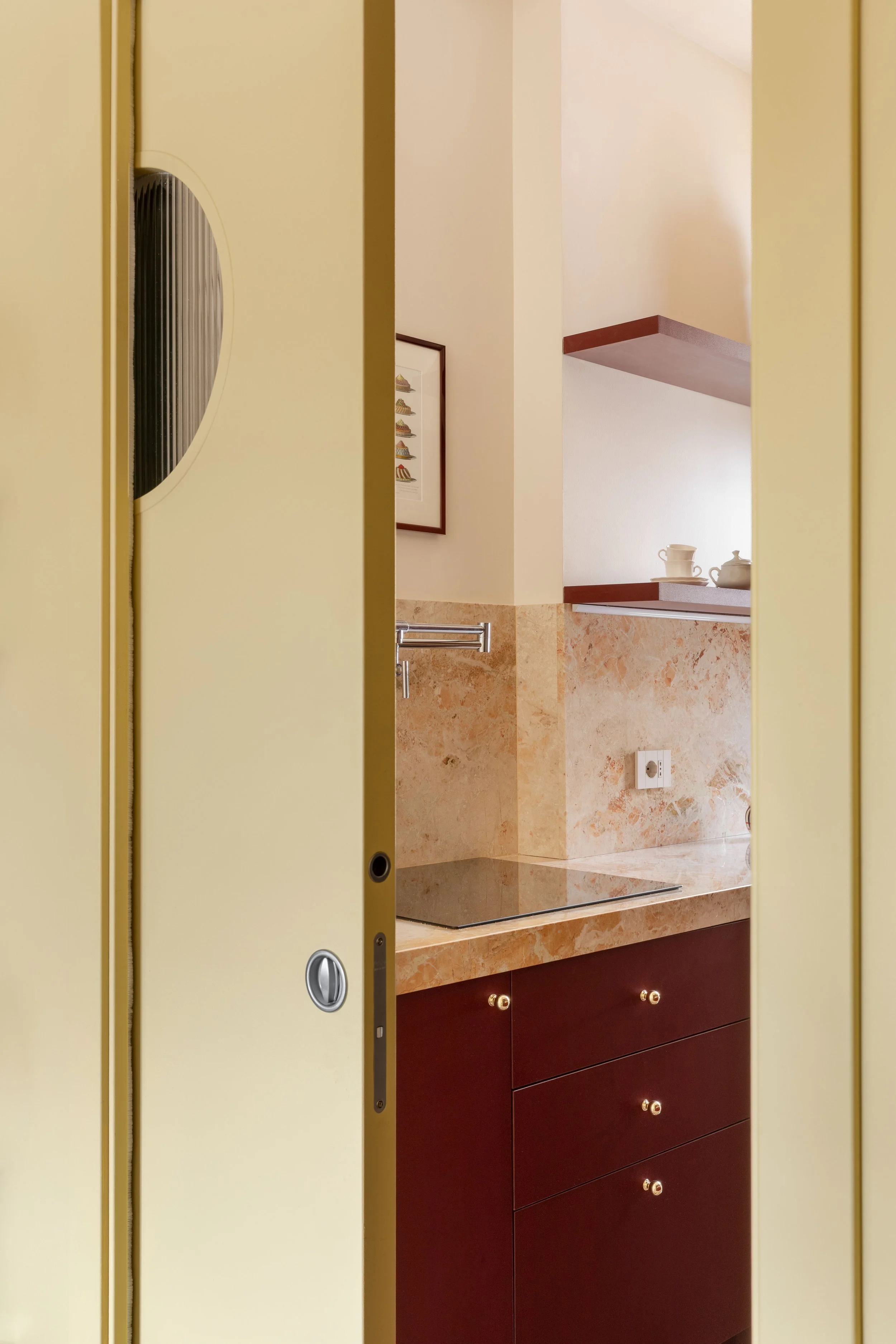Vista parziale di una cucina con ante di legno scure, lavabo e scaffali con tazze e ciotole, visto attraverso una porta gialla.