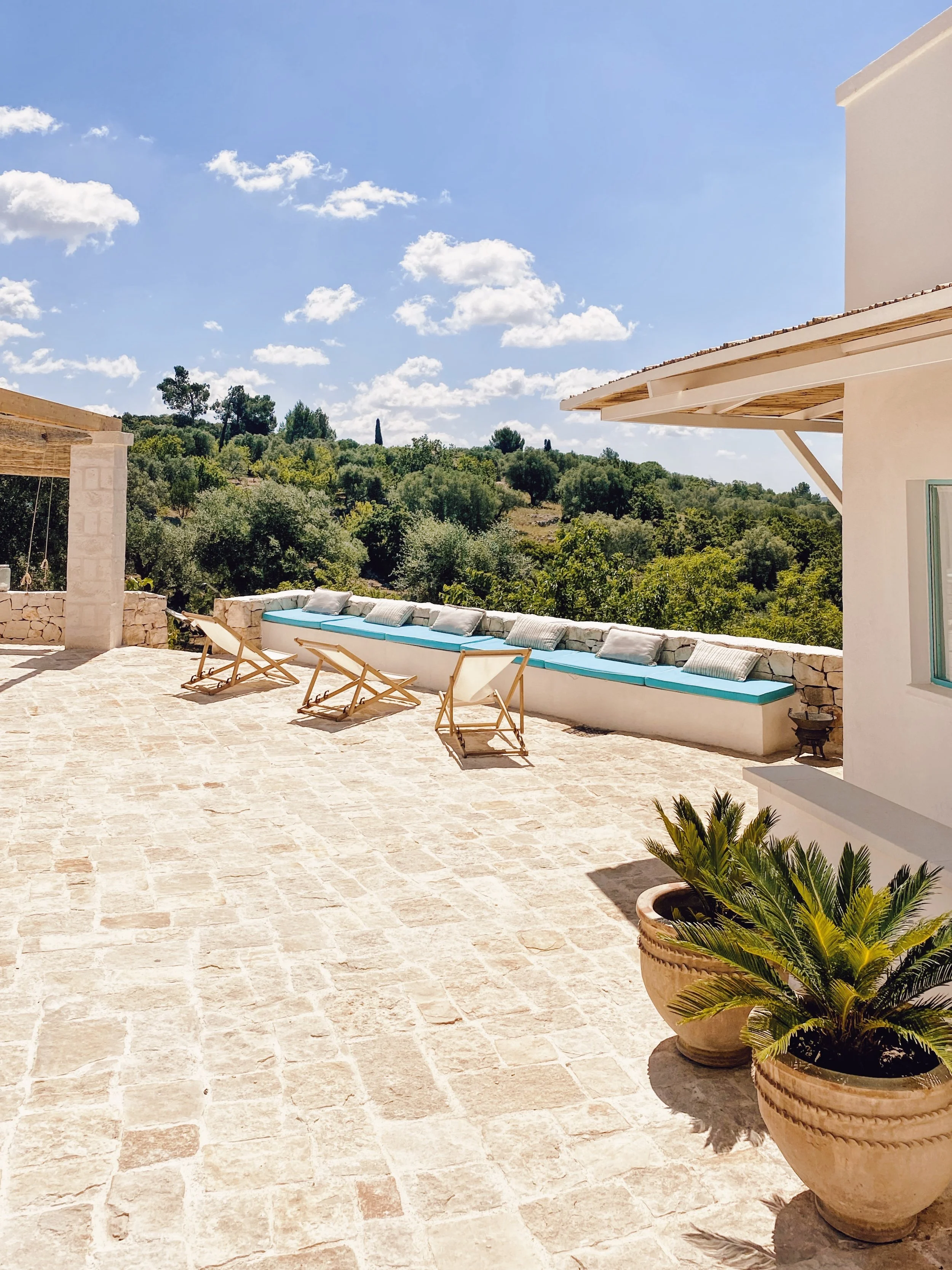 Terrazza con sedie a sdraio, cuscini, una panchina lunga con cuscini, piante in vasi e vista sulla natura con alberi e cielo con nuvole